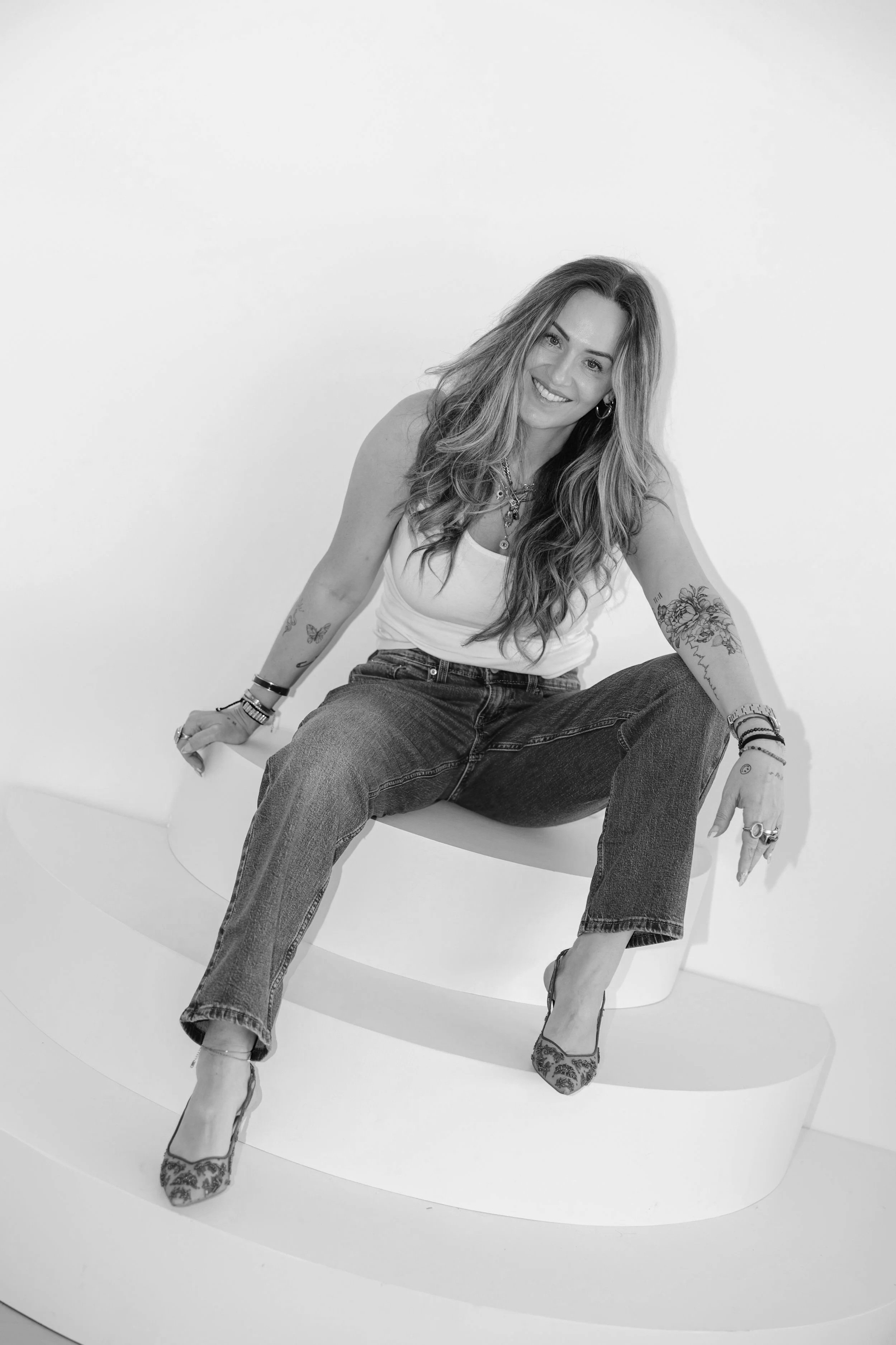 Black and white photo of a smiling woman with long wavy hair, sitting on a modern white chair against a plain background. She is wearing a tank top and jeans, with tattoos on her arms, jewelry including earrings, necklaces, bracelets, and rings, and high heels.