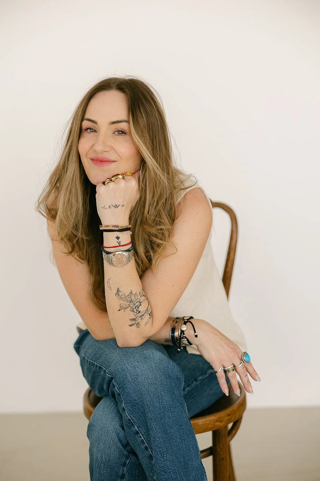 A woman with long, wavy hair and tattoos sits on a wooden chair, smiling and resting her chin on her hand, against a plain off-white background.