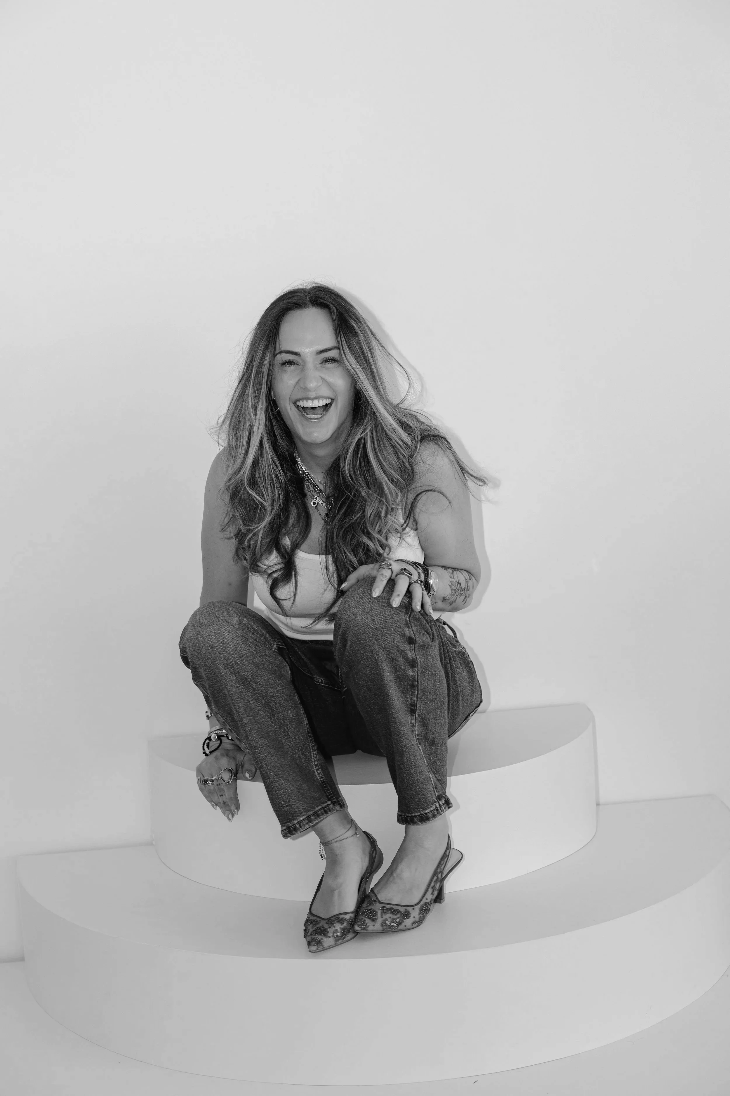 Black and white photo of a woman sitting on a curved seat against a blank wall, laughing with her mouth open and her eyes squinting, wearing a sleeveless top, jeans, high heels, and multiple jewelry accessories.