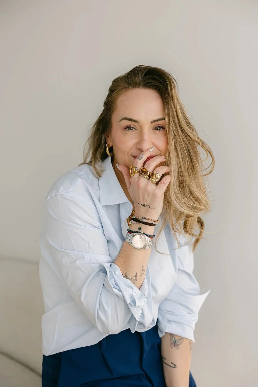 A woman with wavy, light brown hair and multiple tattoos, wearing a white shirt with rolled-up sleeves, blue pants, and an array of jewelry, including gold rings, earrings, bracelets, and a silver watch, sitting against a neutral background.