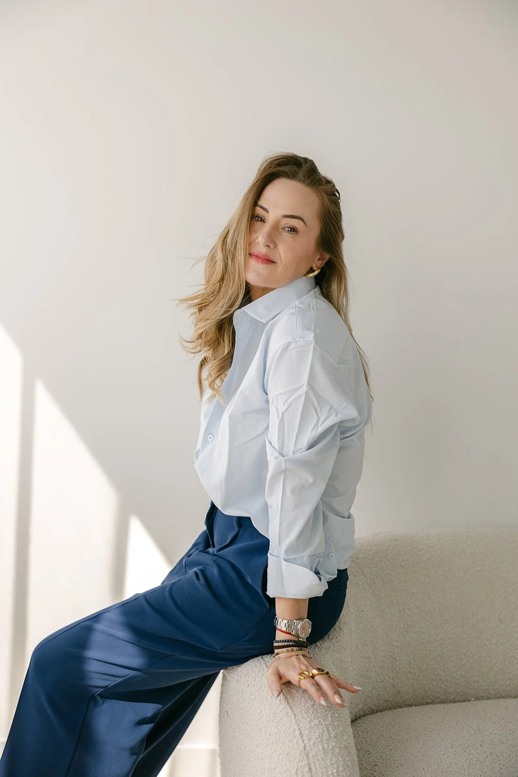 A woman with long, wavy blonde hair is sitting on a textured beige chair, wearing a white button-up shirt with rolled-up sleeves and dark blue pants. She has a relaxed expression with a slight smile, gold earrings, a silver watch, and multiple bracelets and rings. The background is plain and light-colored with natural light casting soft shadows.