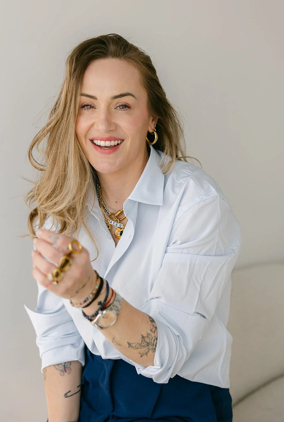 A woman with long blonde hair, wearing multiple jewelry pieces and a white shirt, smiling and looking at the camera.