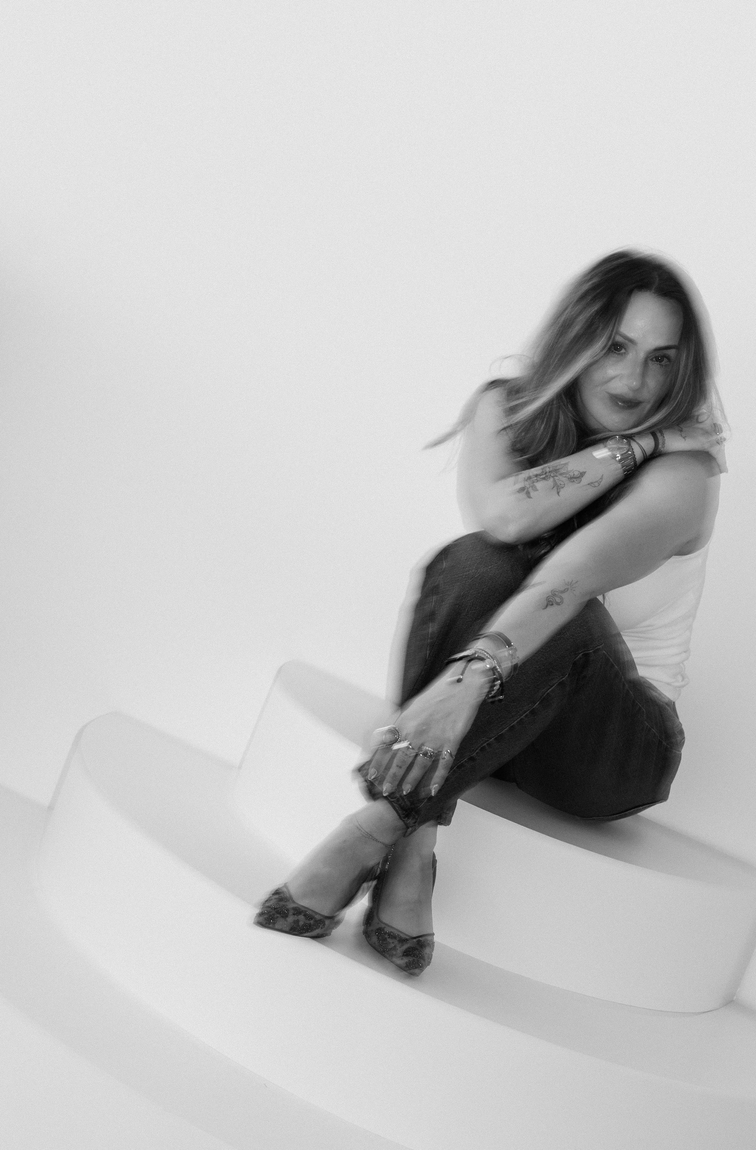 Black and white photo of a woman sitting on a curved bench with her arms crossed over her knees, looking at the camera, with tattoos on her right arm and wearing high heels.