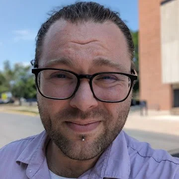 Close-up of a man with glasses, a beard piercing, and slicked-back hair, outdoors on a sunny day.