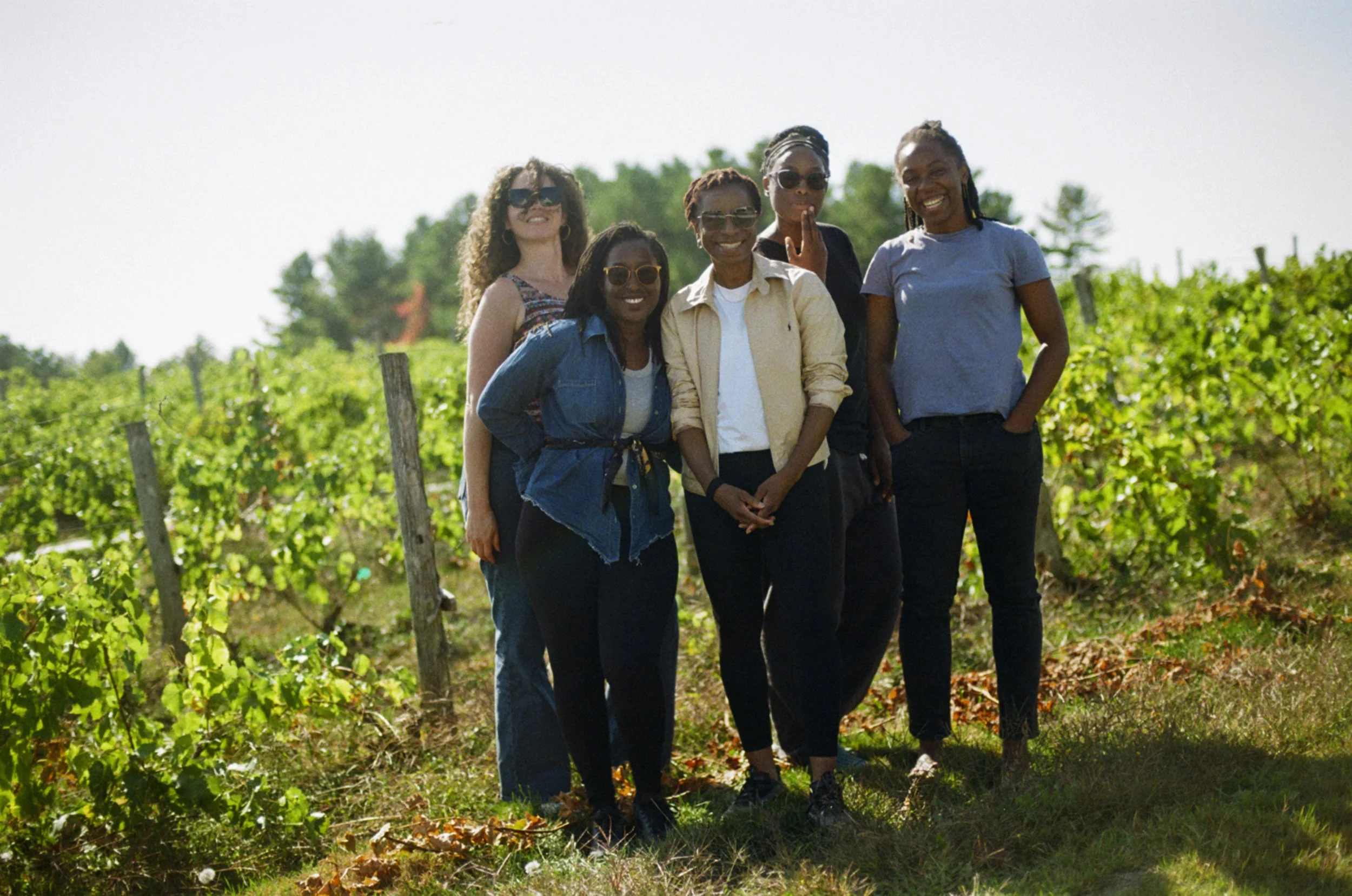 Group of six women standing together in a vineyard, smiling on a sunny day.