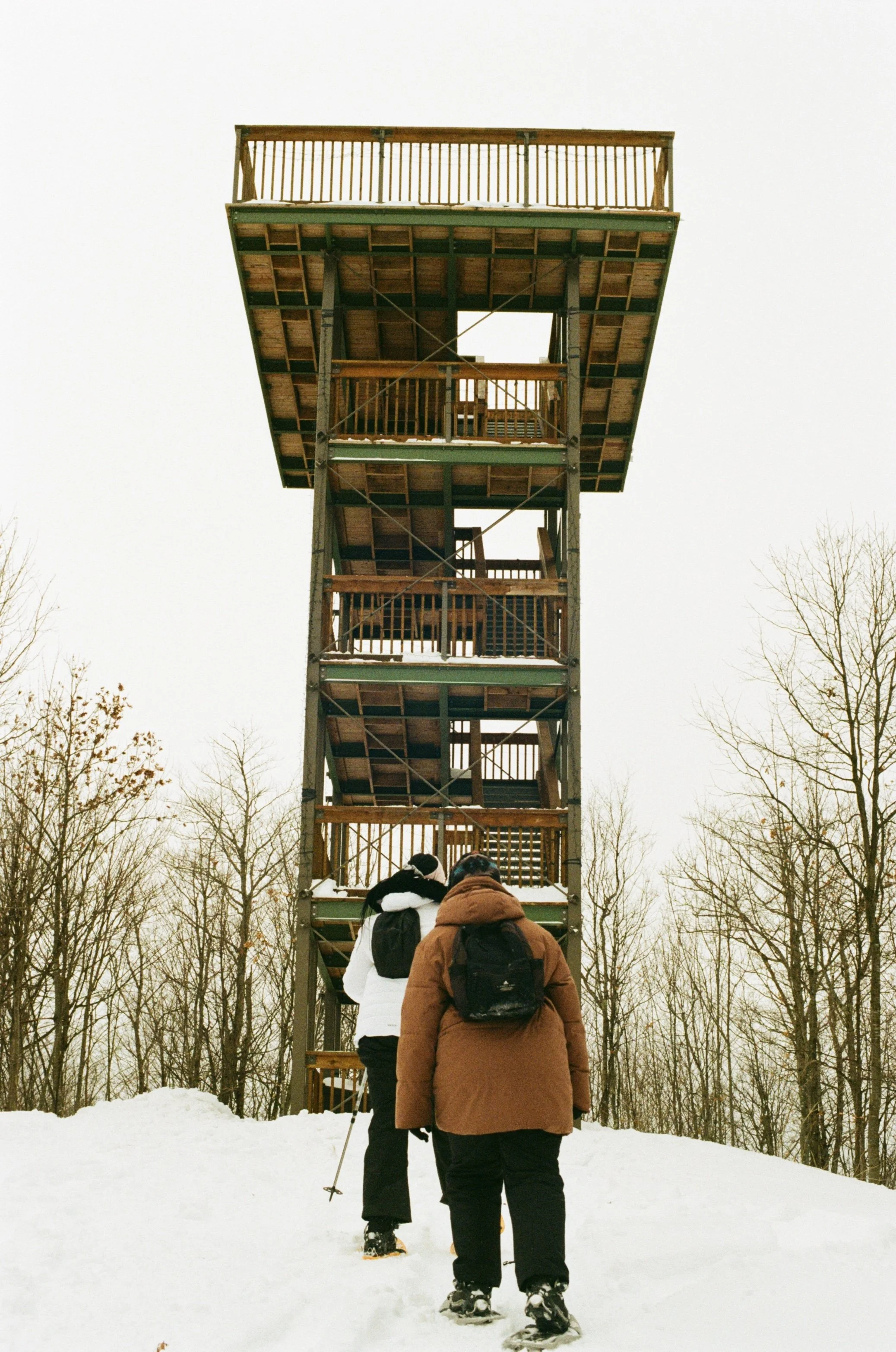 Two people with backpacks and snowshoes walking towards a tall wooden observation tower in a snowy forest during winter.