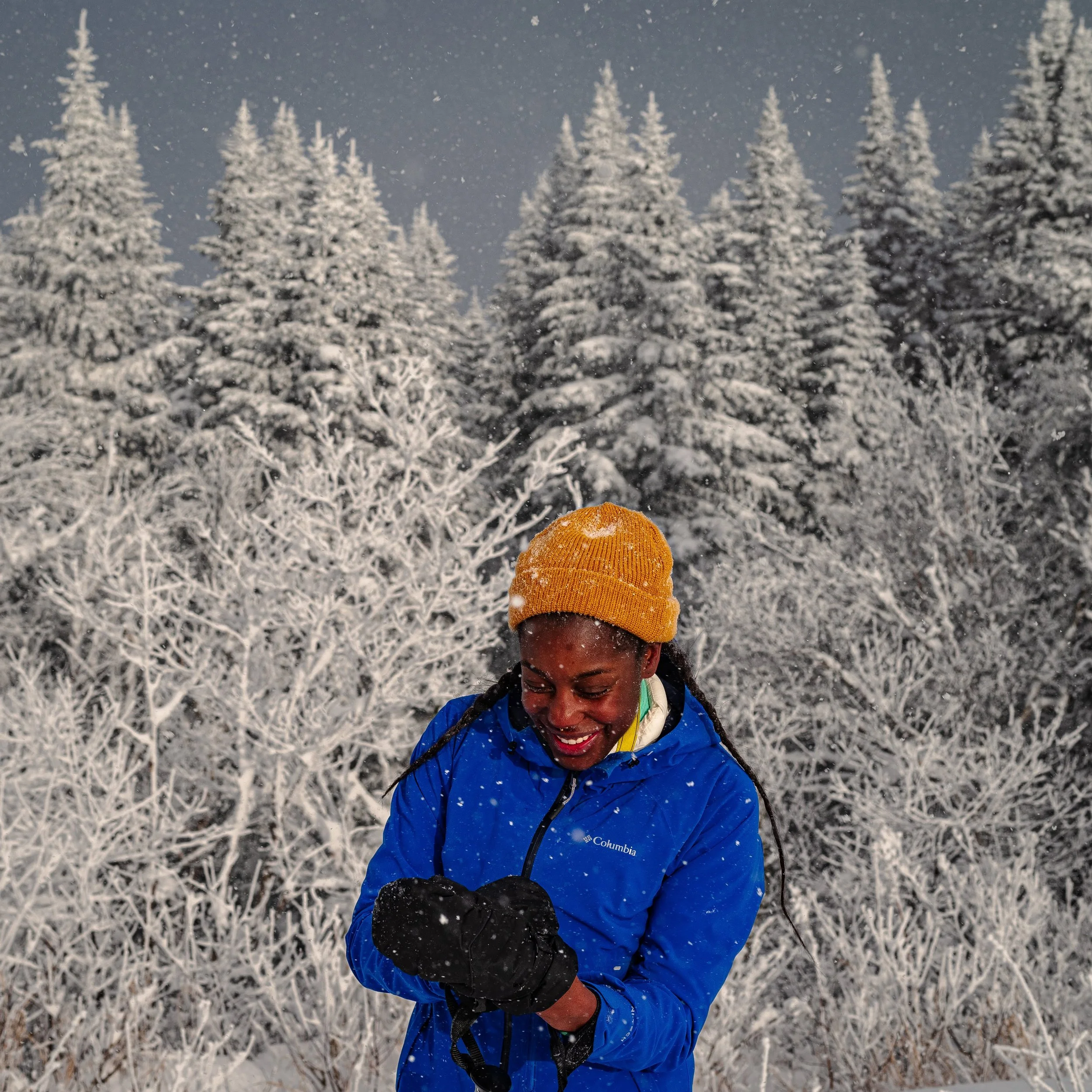 A woman dressed in a blue Columbia jacket and yellow beanie smiling and looking down while holding her gloves in a snowy forest with snow-covered trees in the background.