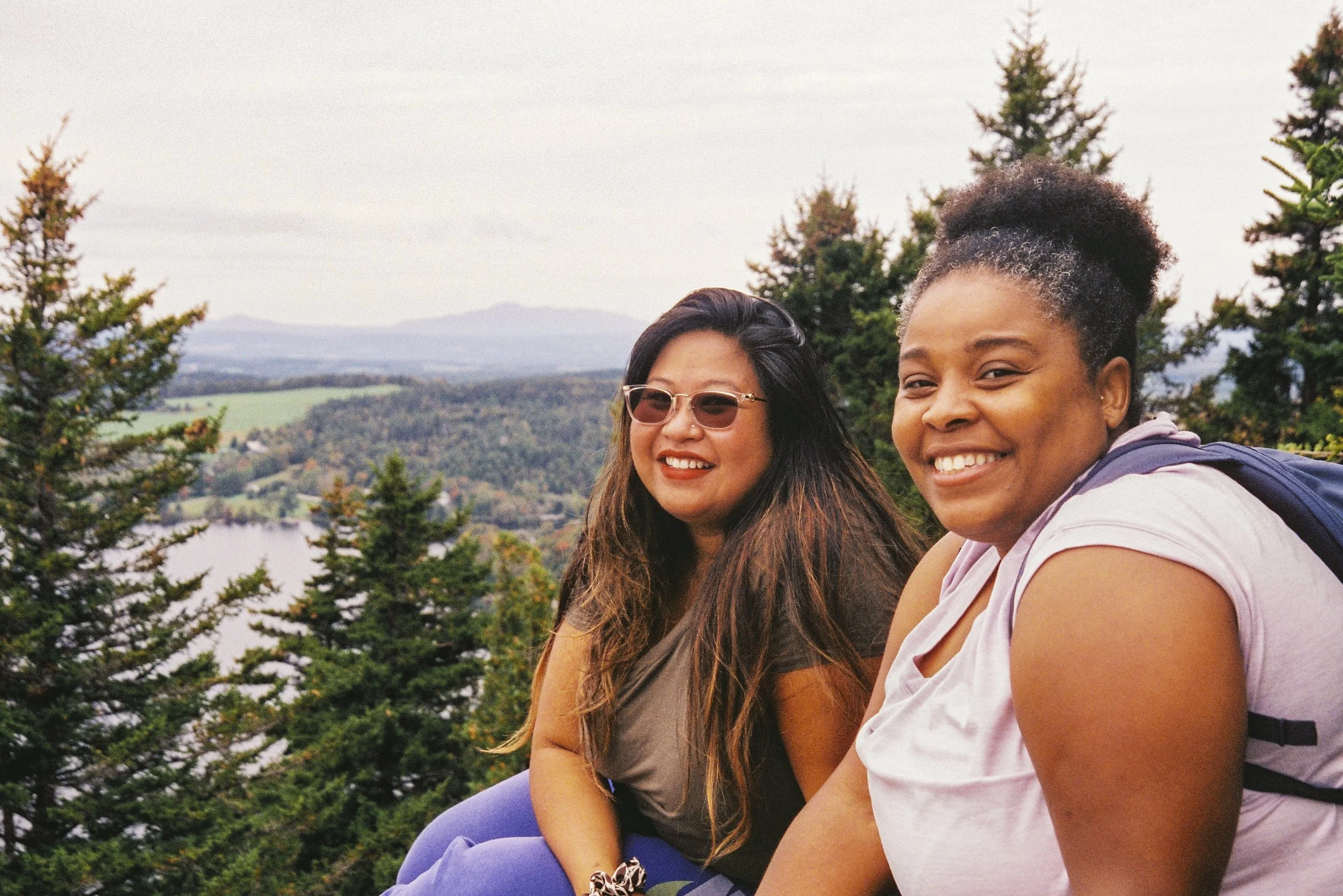 Two women sitting outdoors among trees, smiling, with a scenic view of a lake, rolling hills, and mountains in the background.