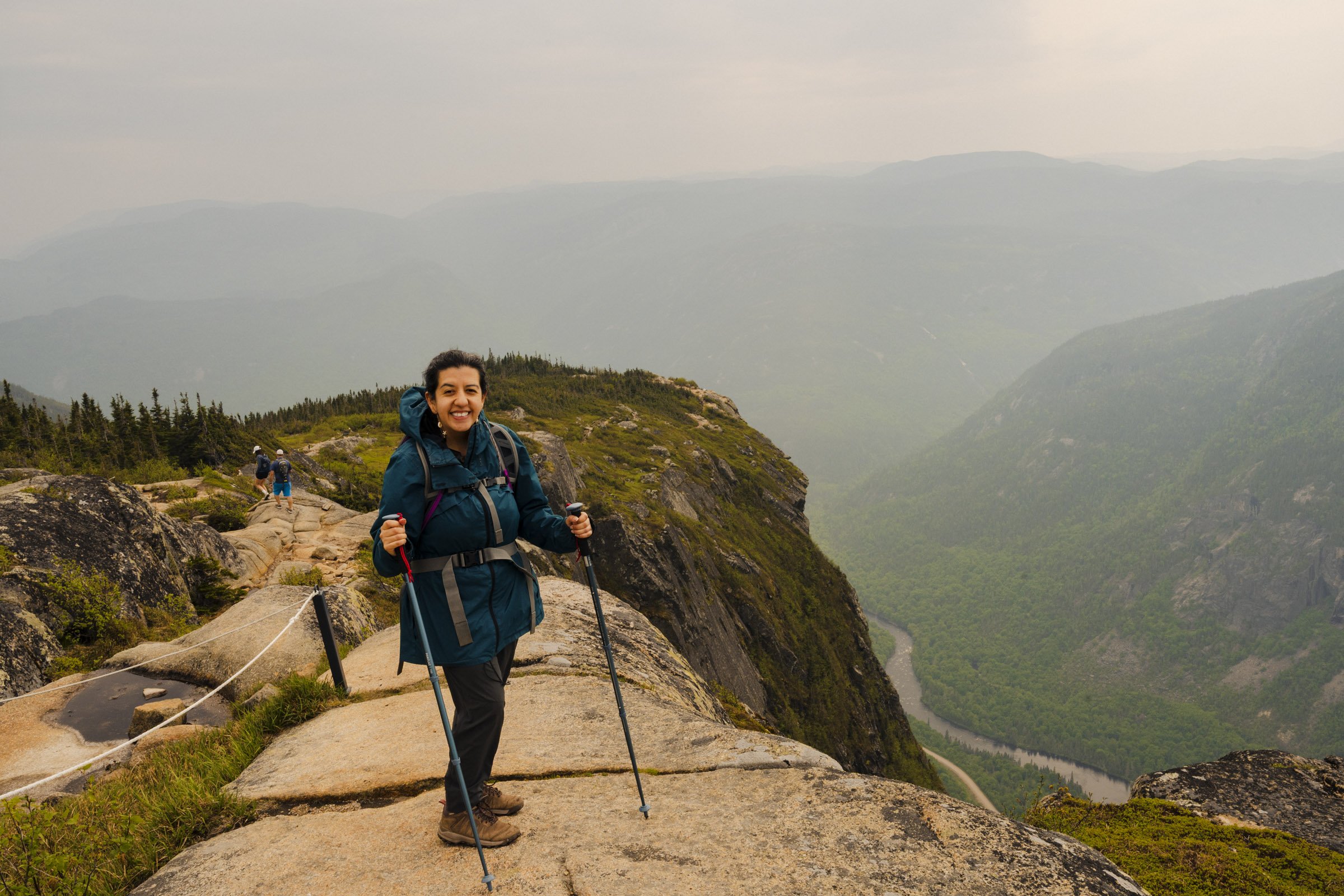 Woman in hiking gear smiling at the camera on a mountain trail with a scenic view of mountains, a river, and forest in the background.