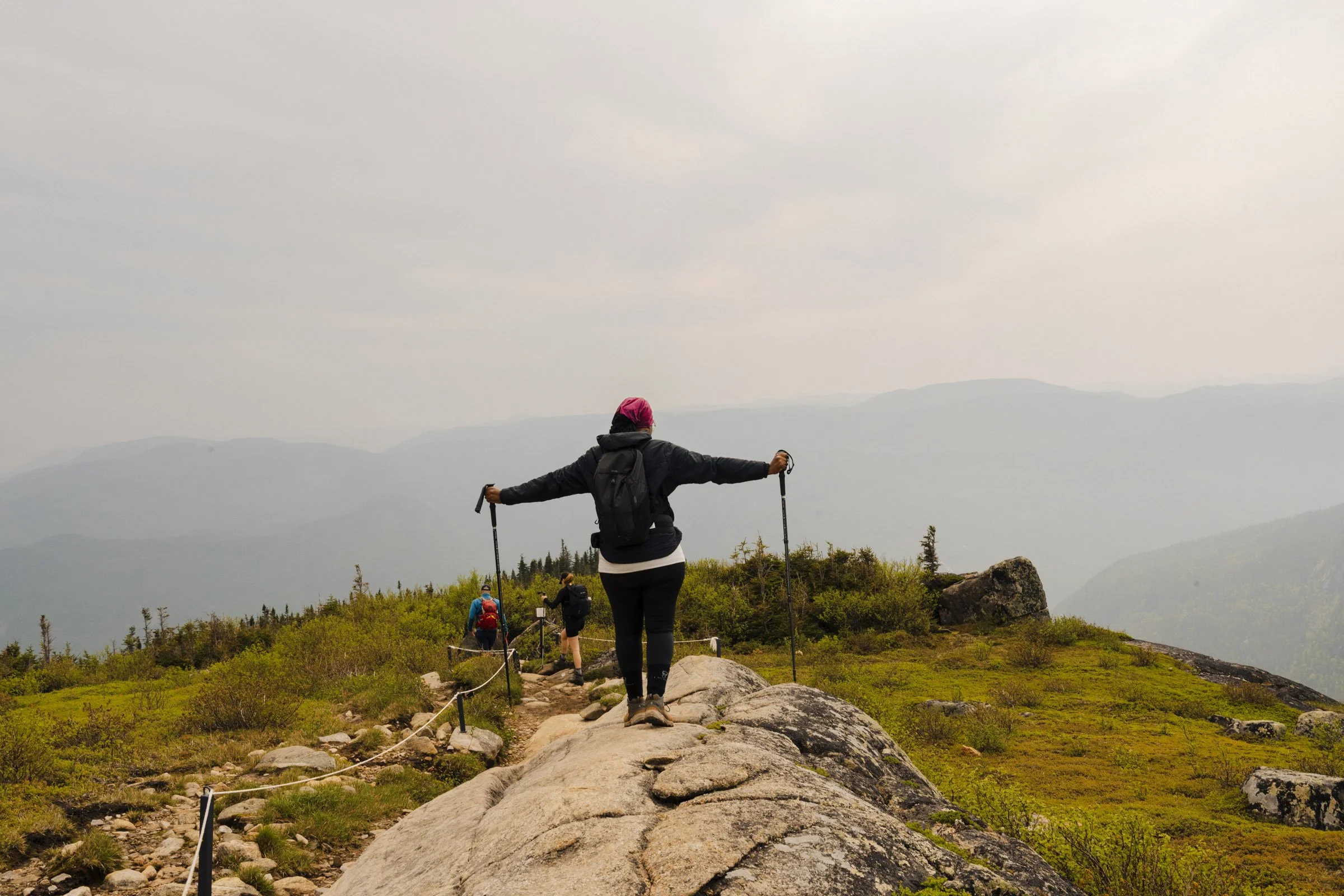 Person in black jacket and pink head covering standing on a mountain trail with arms outstretched holding trekking poles, overlooking a foggy mountain landscape with other hikers ahead.