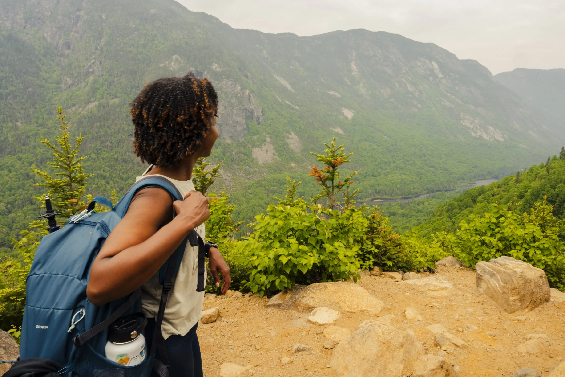 A woman with curly hair wearing a backpack and looking out at a mountainous landscape with green trees and a river