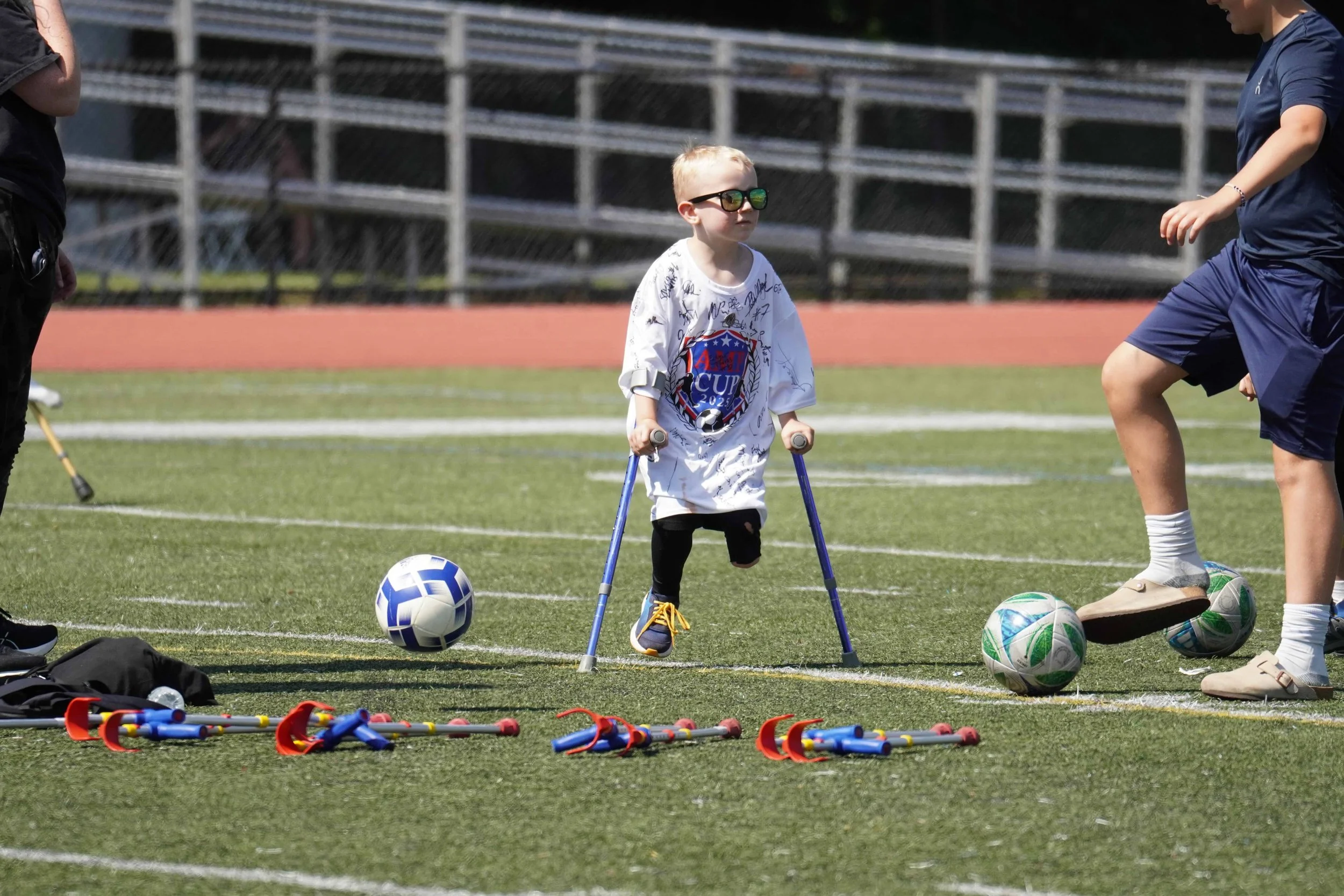 Young boy with sunglasses using crutches on a soccer field, surrounded by soccer balls, with two people nearby, some sports equipment on the ground.