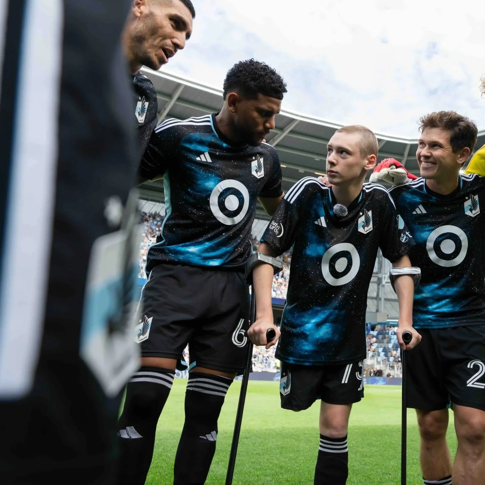 A group of soccer players with disabilities, wearing black jerseys with the Target logo, standing in a huddle on a soccer field, listening attentively.