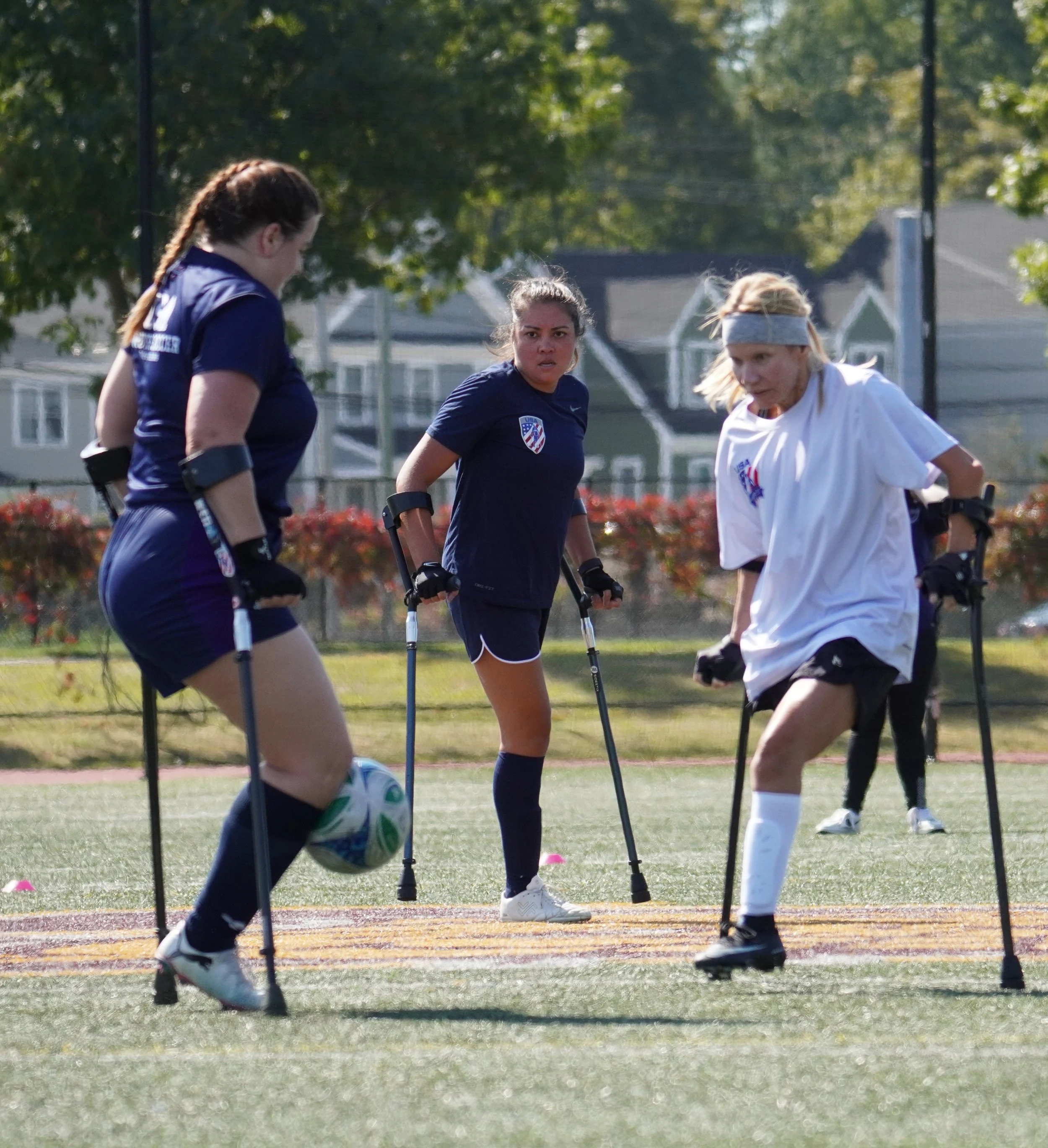 Three women playing soccer on a field, two wearing dark uniforms and one wearing a white uniform, with red cones on the field and hair tied back, practicing with focus.