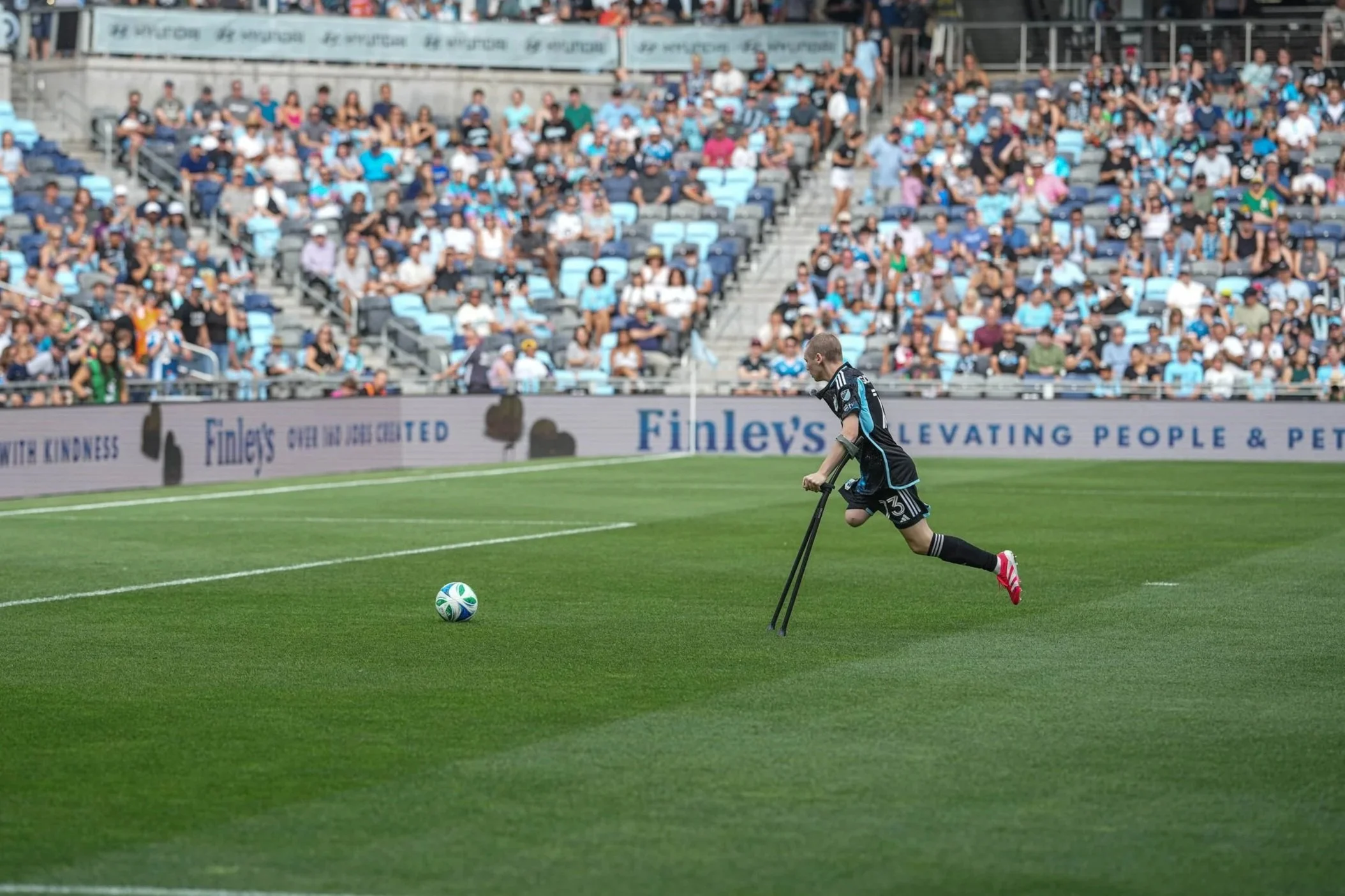 A soccer player with a disability using crutches chases a soccer ball on a green field during a match, with a crowd of spectators in the stands.