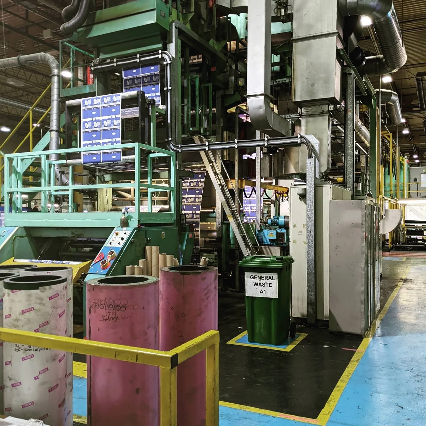 Industrial manufacturing machine with recycled paper rolls, a green waste bin labeled 'GENERAL WASTE,' and interconnected machinery in a factory setting.