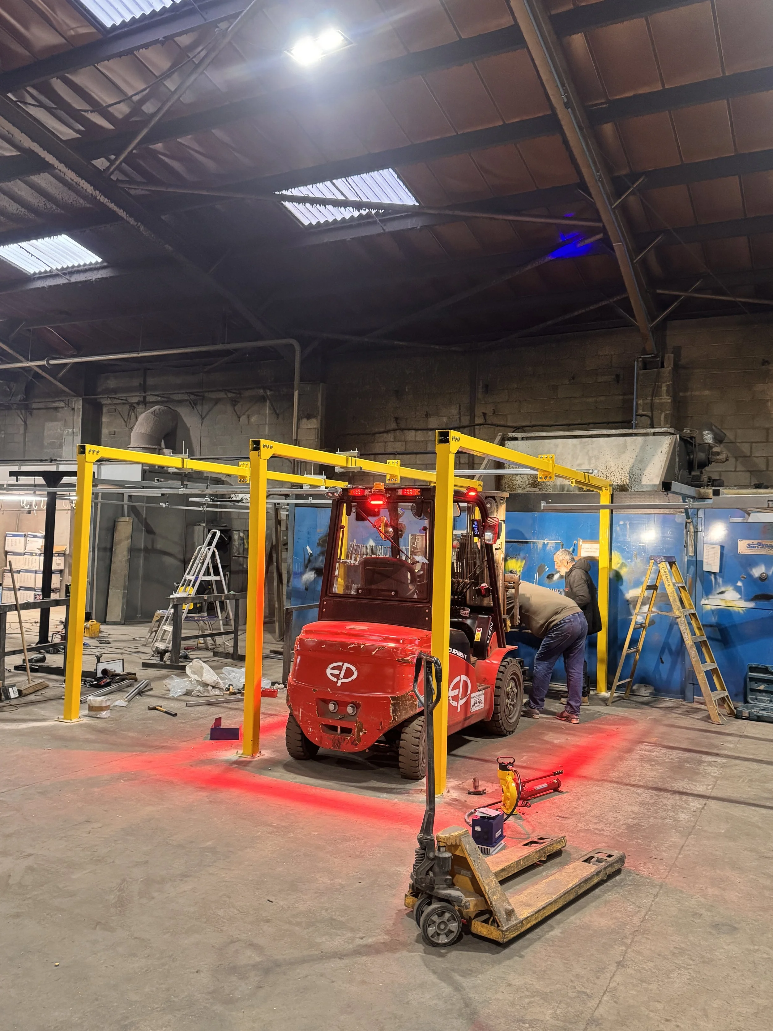 Construction workers are working inside a warehouse or factory. There is a red forklift in the center with two workers on the right side using tools near a blue wall. Various construction equipment and tools are scattered on the floor, and the ceiling has metallic beams and skylights.