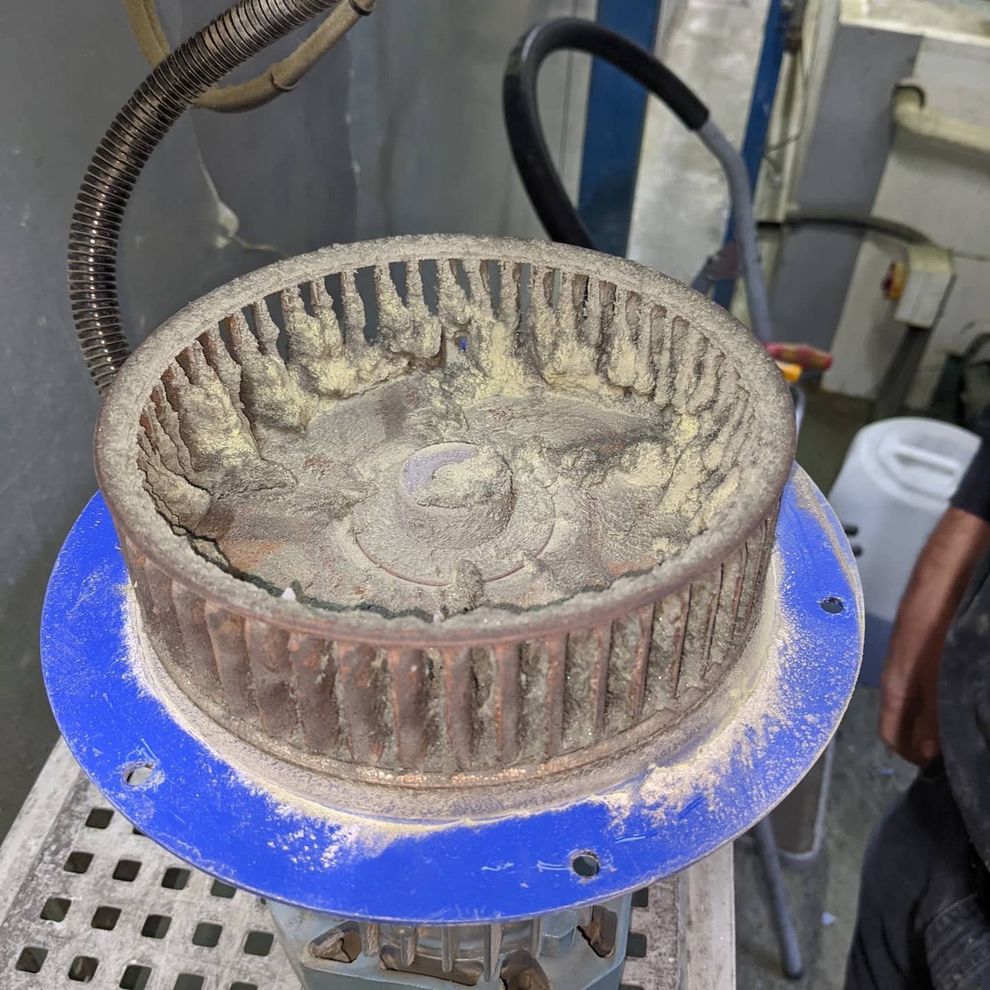 A dusty cylindrical air filter with pleated filter material inside, placed on a blue circular base, in a workshop environment.