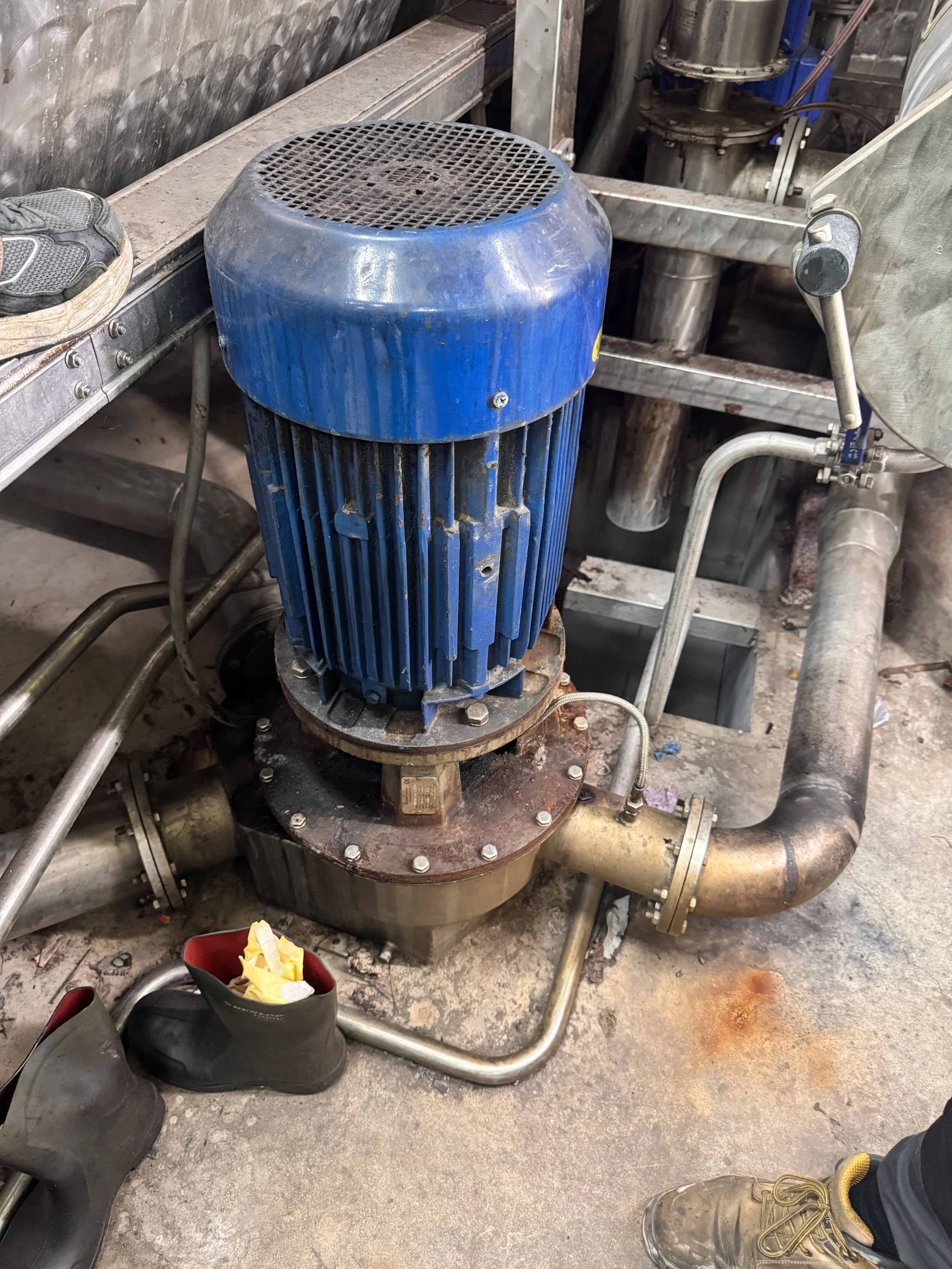 Industrial machinery, a blue electric motor mounted on a rusty flange connected to pipes and metal framework in a factory setting.
