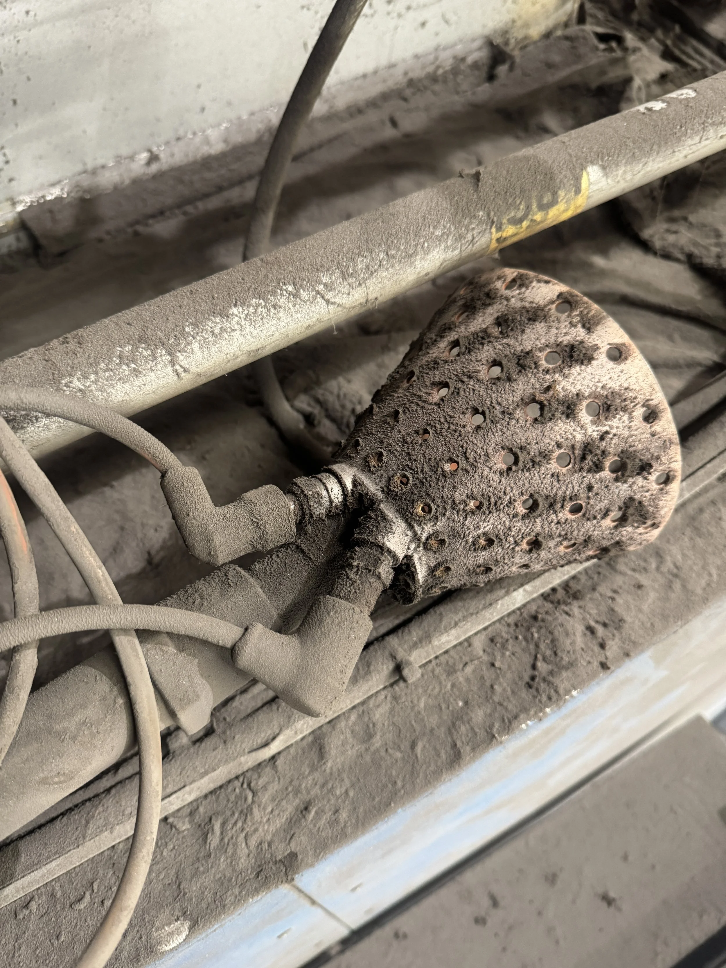 Close-up of dusty, dirty electrical and plumbing components behind a wall or panel, including a perforated metal plate, a pipe, and wiring.