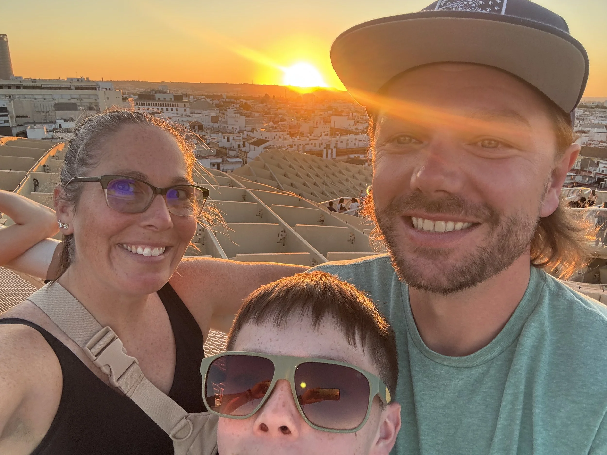 A smiling family of three taking a selfie during sunset with a cityscape background.