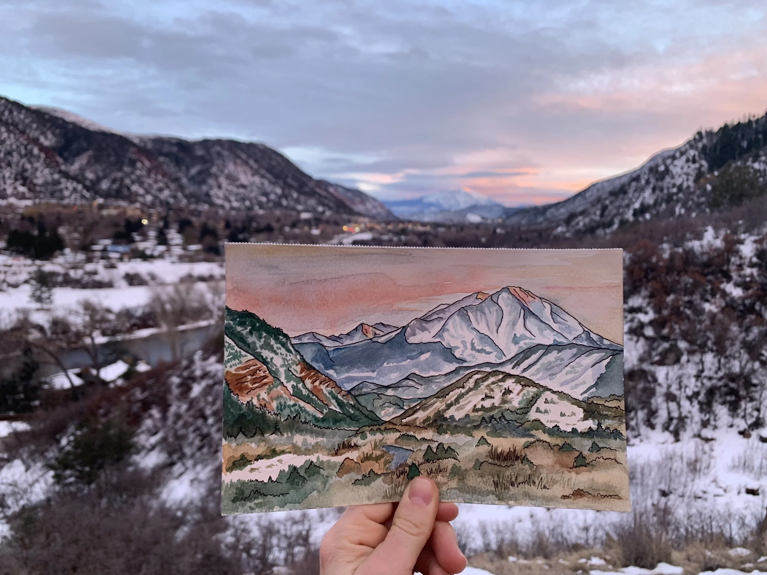 Person holding a watercolor painting of snow-capped mountains against a sunset sky in front of a real snowy mountain landscape.