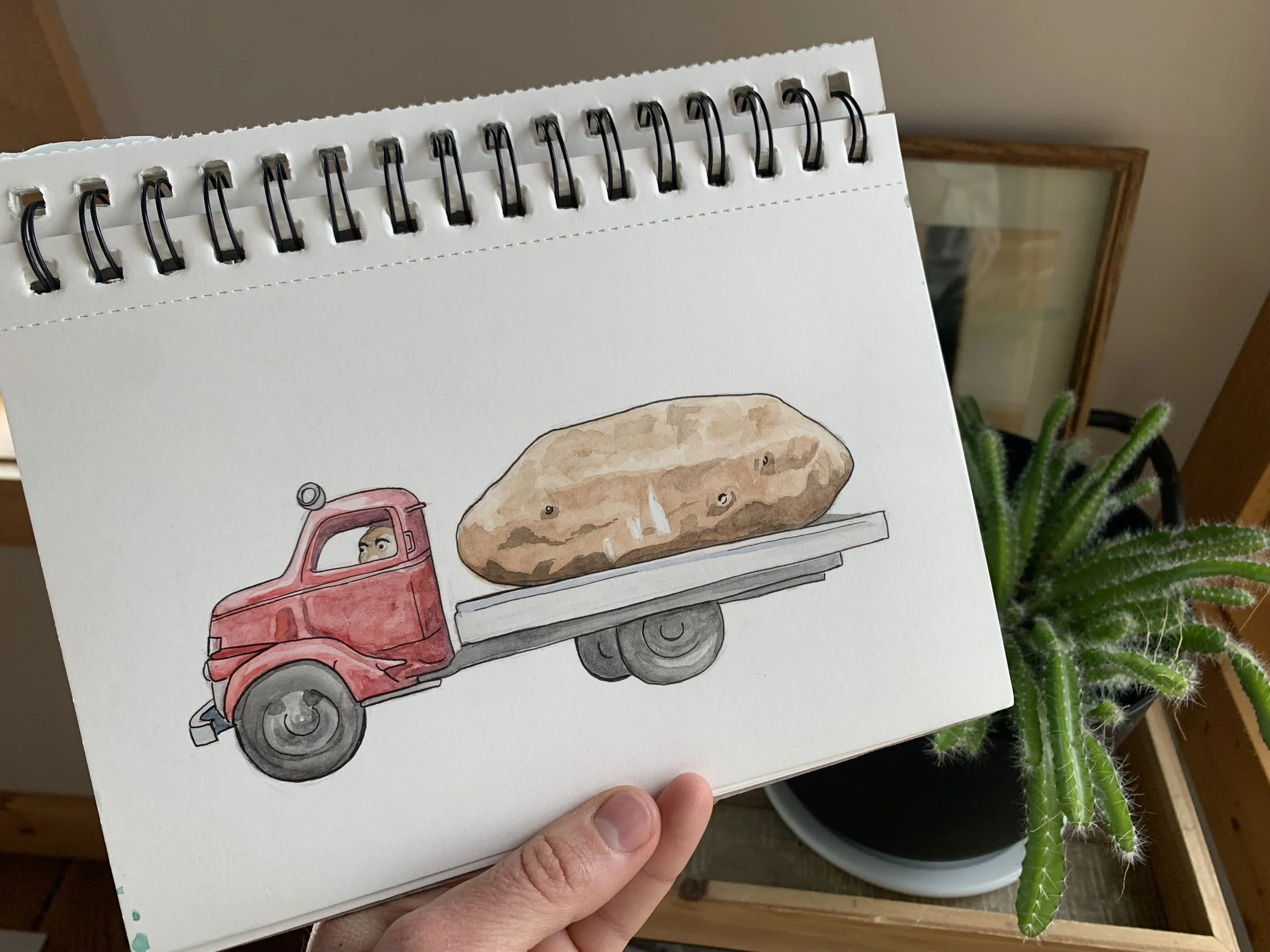 Hand holding a sketchbook with a watercolor drawing of a red flatbed truck carrying a large rock, with a plant visible in the background.