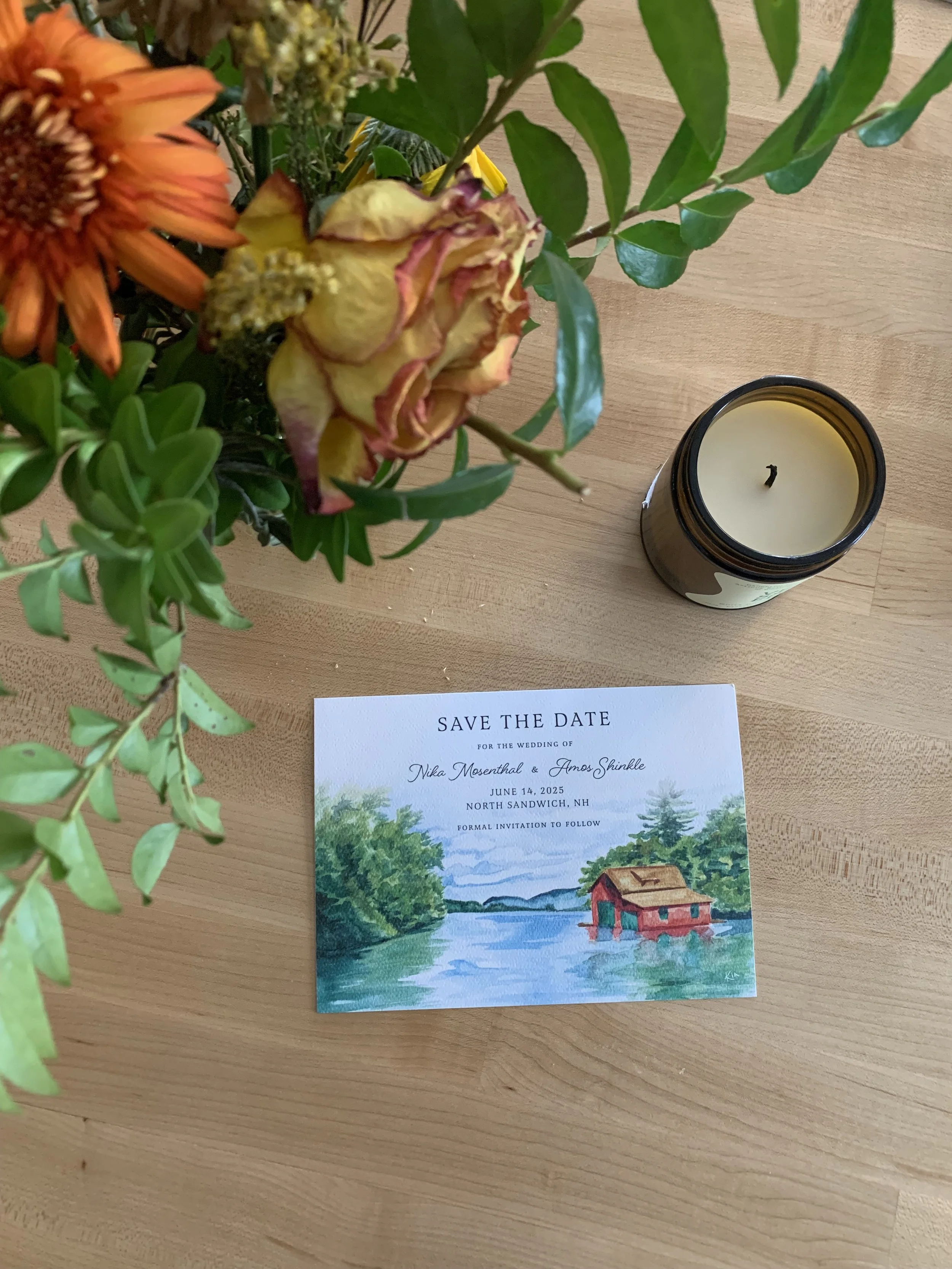 A table with a bouquet of flowers, a black candle jar, and a watercolor-style wedding save-the-date card for Nika Mosenthal and Amos Shinkle, wedding date June 14, 2025, in North Sandwich, NH.