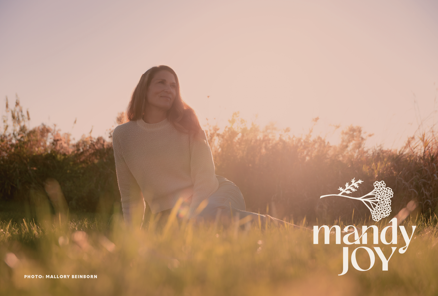 A woman sitting on the grass during sunset, with trees and sunset in the background, and the text 'mandy JOY' with a floral illustration in the lower right corner.