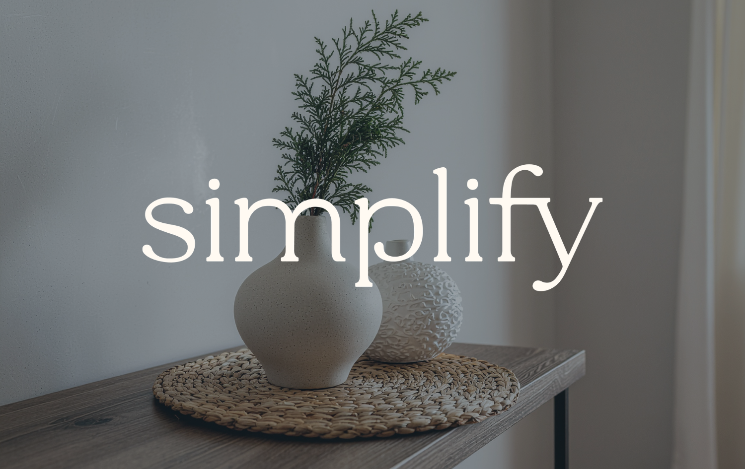A minimalist interior with a wooden table holding a white ceramic vase with greenery and a textured white spherical object, placed on a woven mat. The background is a plain wall with soft lighting. The word 'simplify' is overlaid on the image.