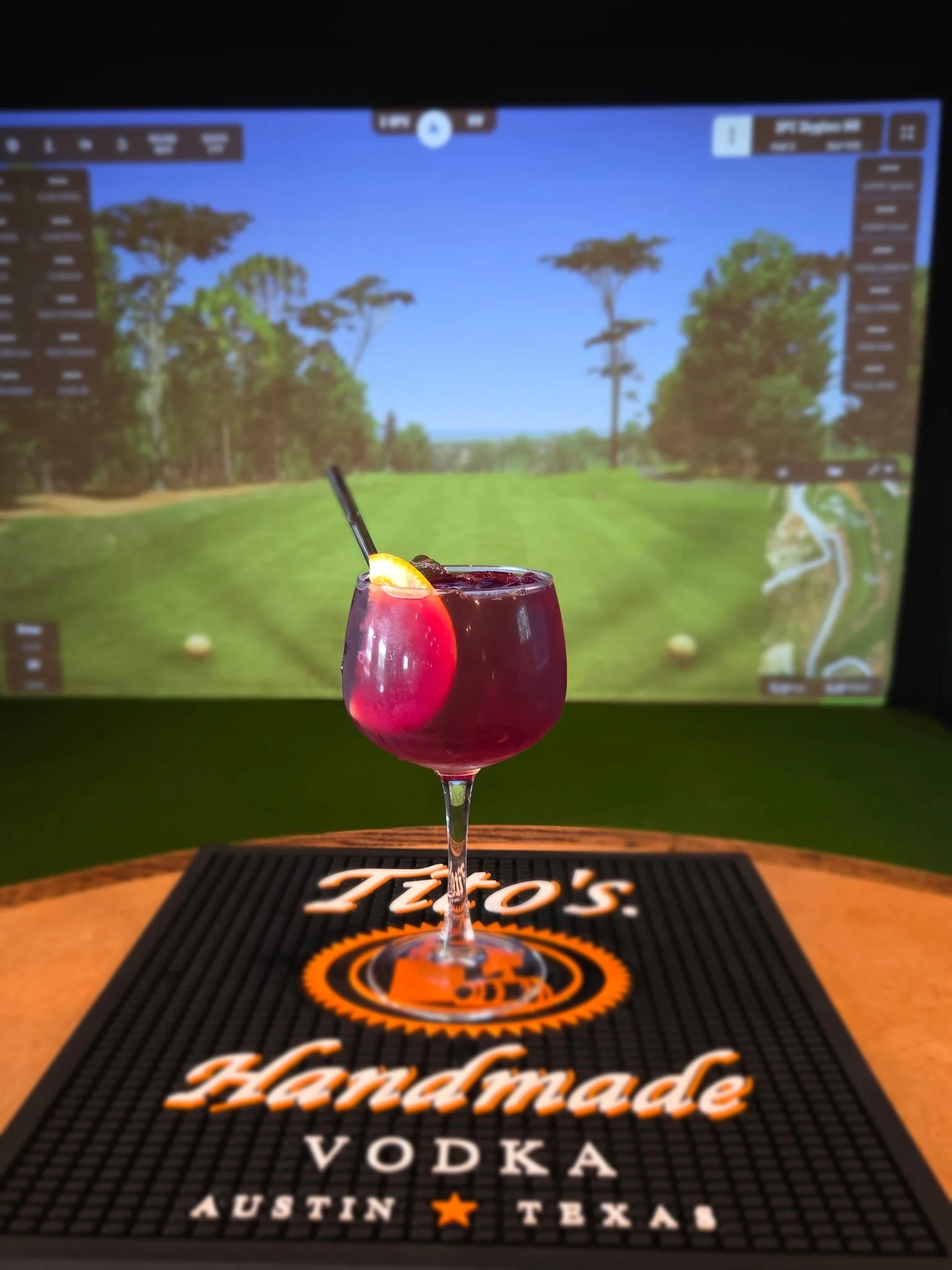 A colorful cocktail glass with a cherry, orange slice, and black straw in front of a virtual golf course backdrop, placed on a wooden surface with a Jack Daniel's branded mat.