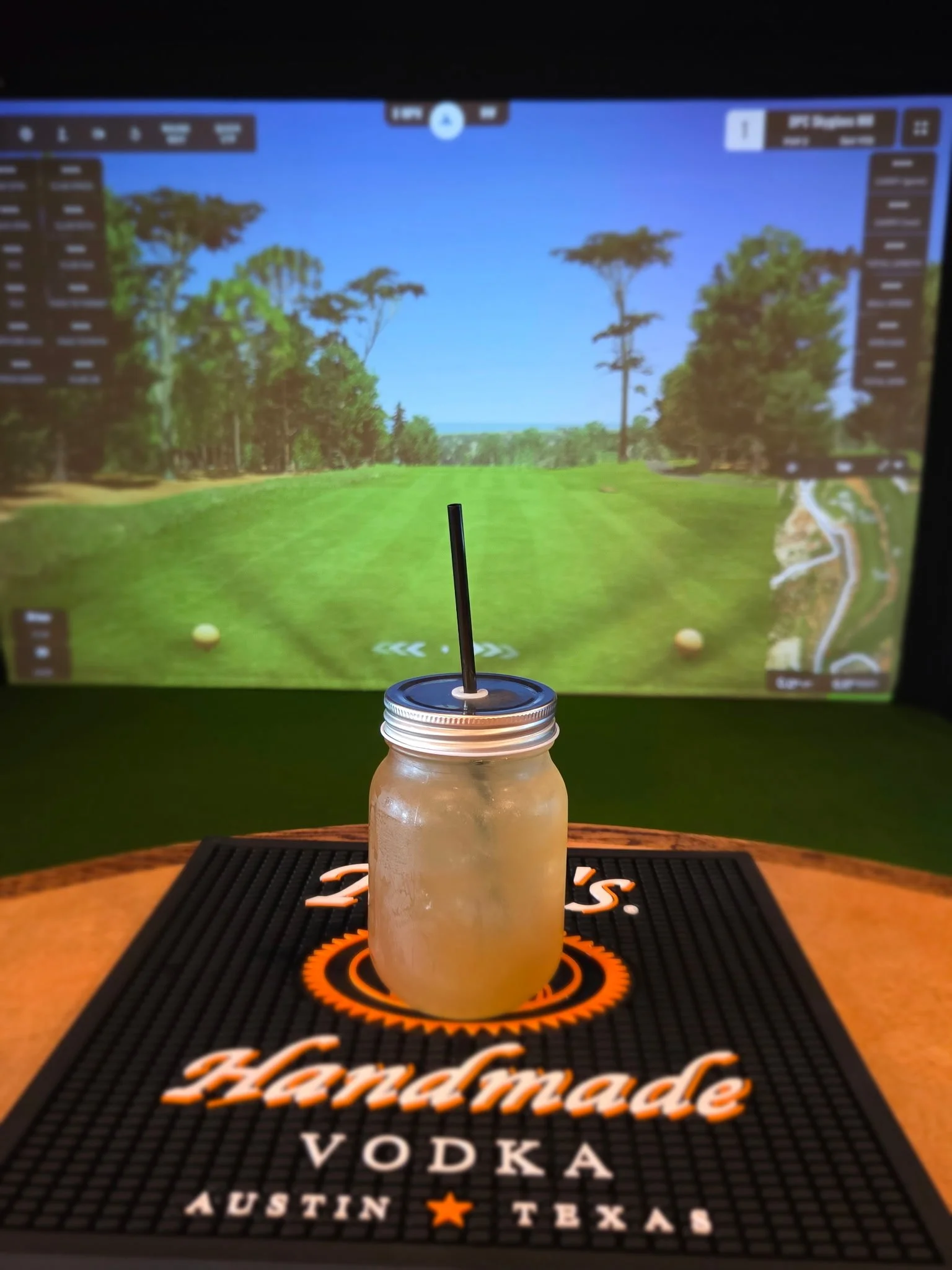 A mason jar with a black straw on a Hardest Vodka mat, with a golf course landscape shown on a screen in the background.