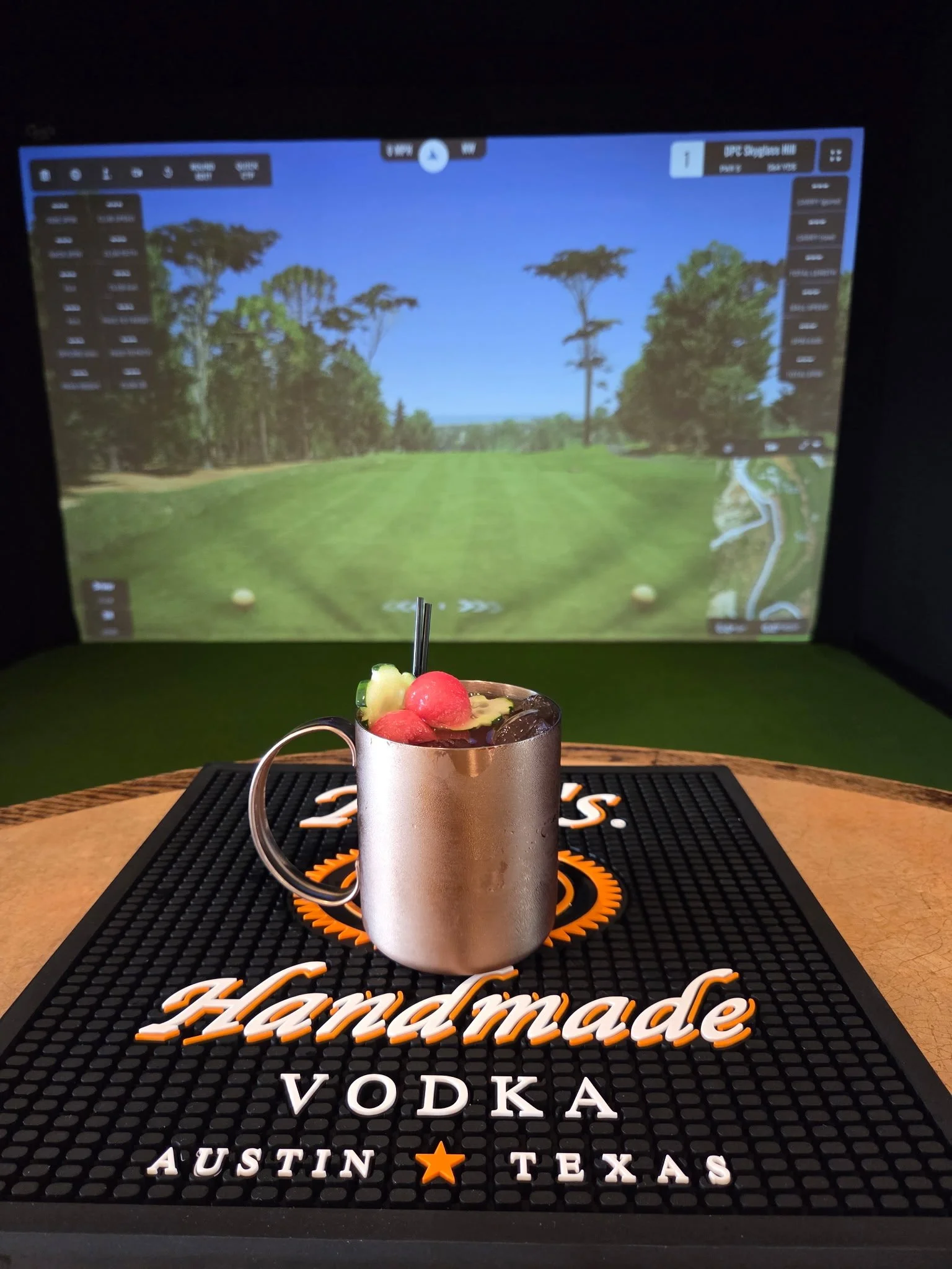 A metal mug containing a colorful fruit cocktail with watermelon, pineapple, and grapes, sitting on a bar mat with the 'Hendmade Vodka' logo on it, in front of a large screen showing a golf course with trees and a blue sky.