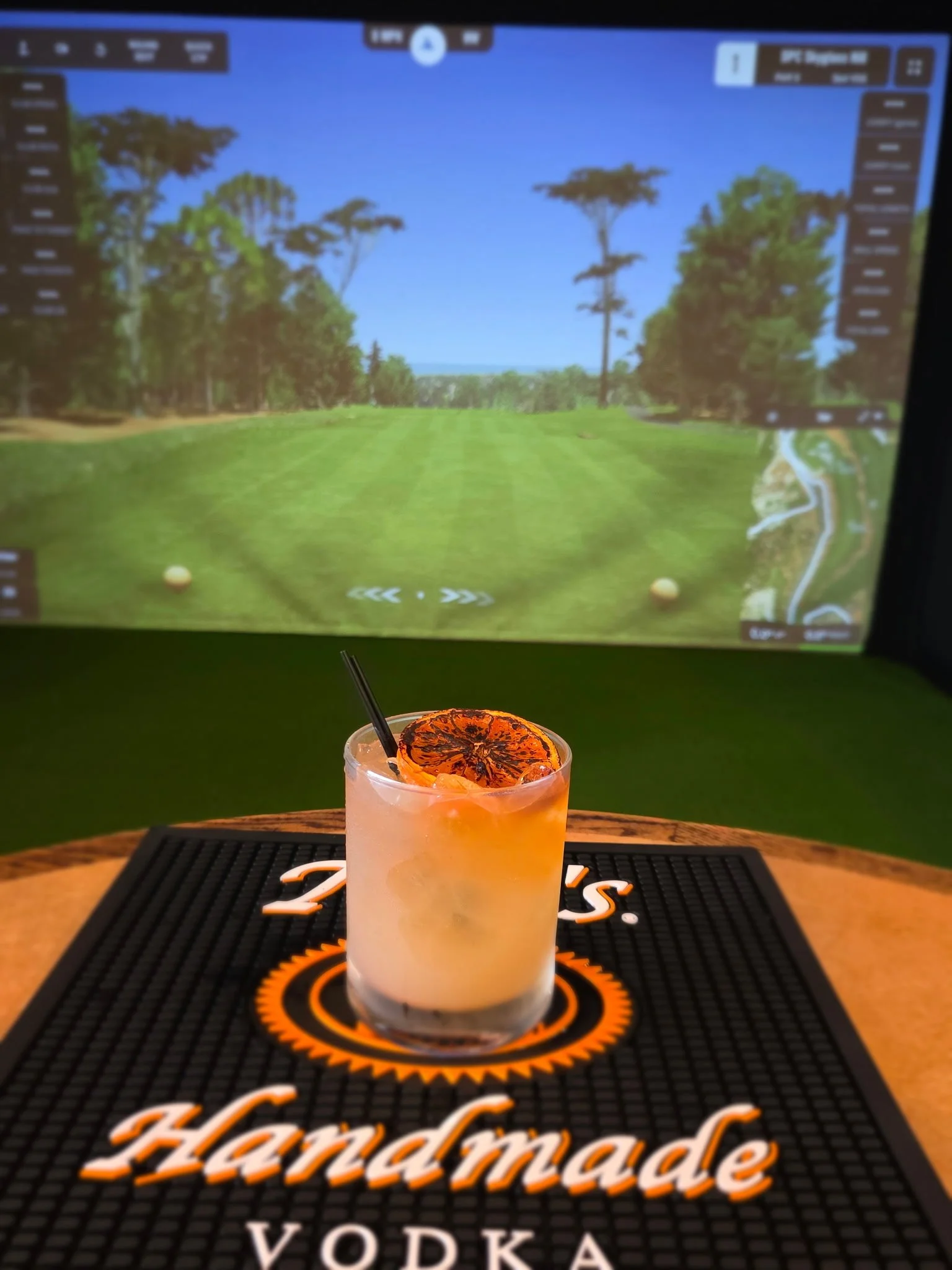 A cocktail topped with a dried orange slice on a bar mat, with a blurred dance floor and a large screen showing nature scenery in the background.