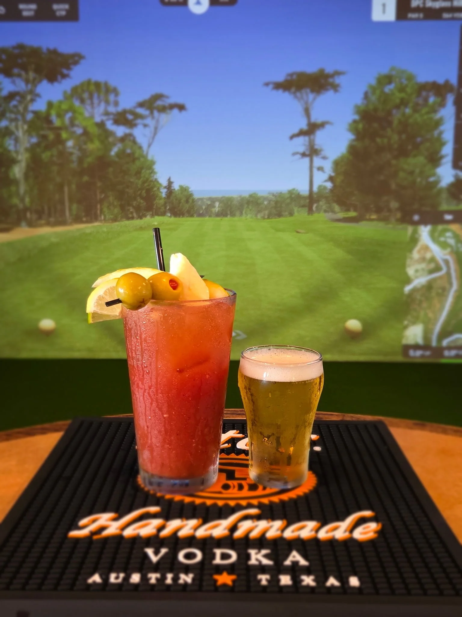 A pink cocktail garnished with lemon and green olives, alongside a smaller glass, set on a bar mat featuring the text 'Hardmaded Vodka Austin Texas'.