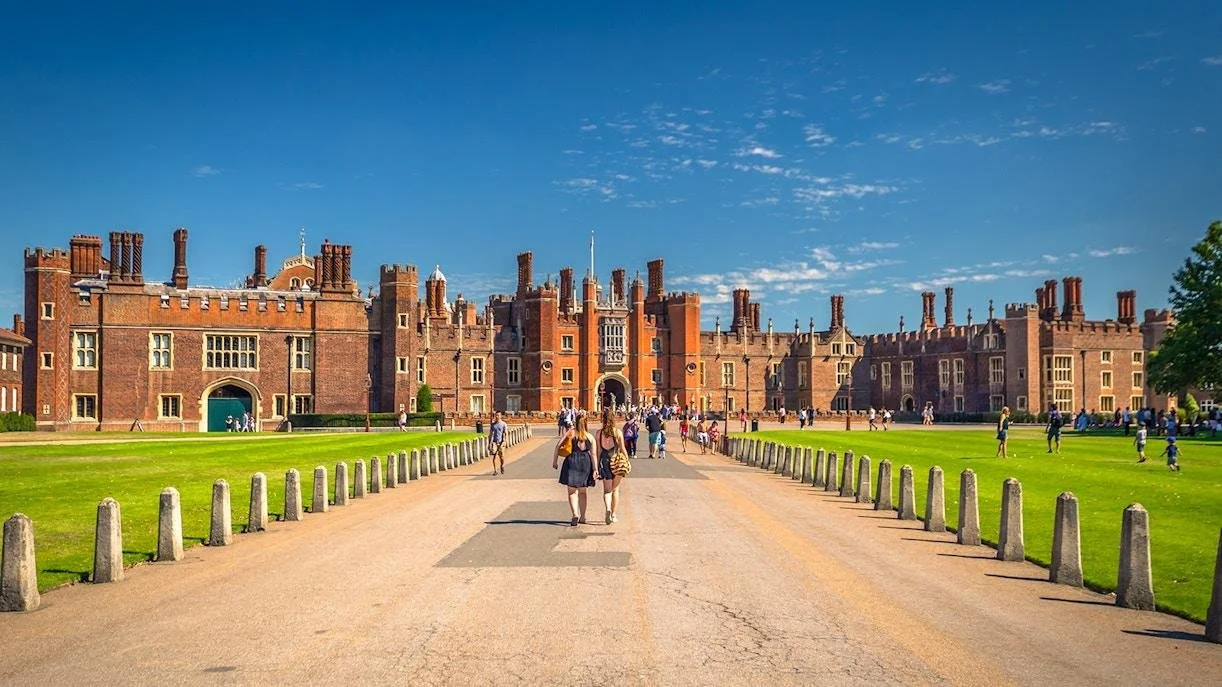 The image depicts a large historic castle with brick walls and numerous chimneys, set against a blue sky with some clouds. The castle is surrounded by a well-maintained green lawn, and there are visitors walking along a wide pathway leading to the castle entrance.