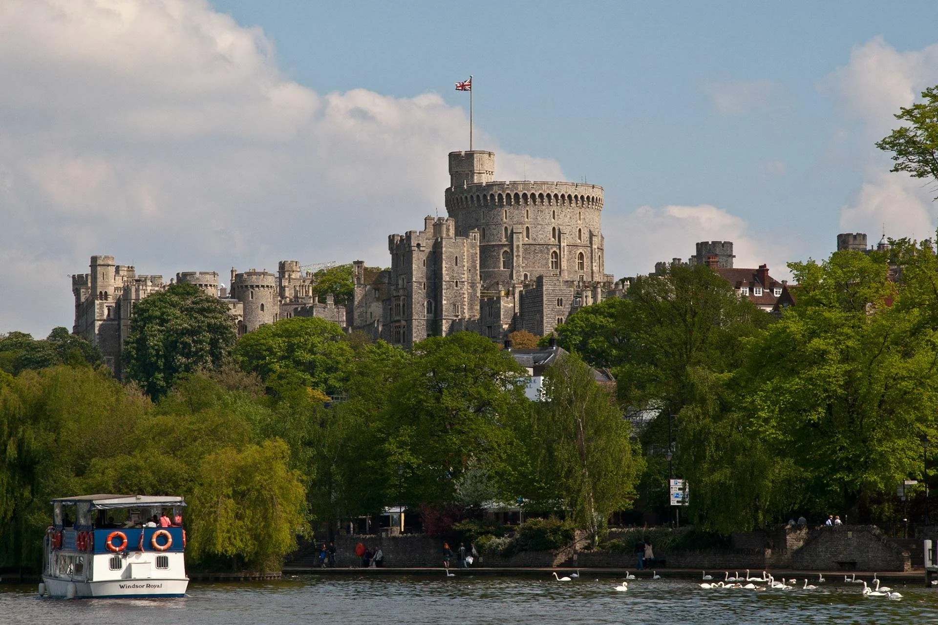 A castle with a tall tower and battlements rising above trees, flying a Union Jack flag, situated on a hill near a river, with boats and swans on the water, and a boat with tourists in the foreground.