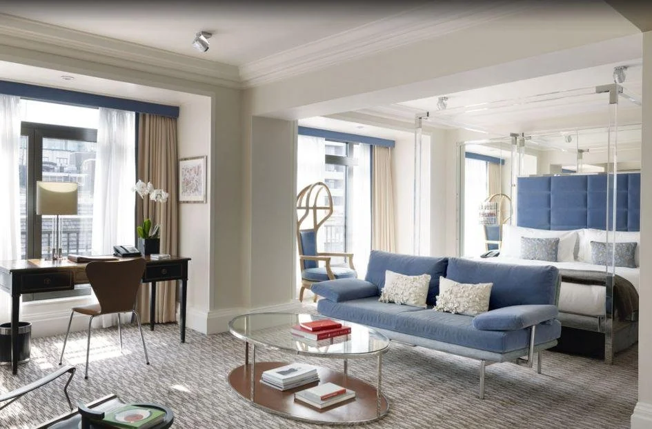 Modern hotel suite with a blue sofa, glass coffee table, and a bedroom with a blue headboard. Large windows with curtains let in natural light.