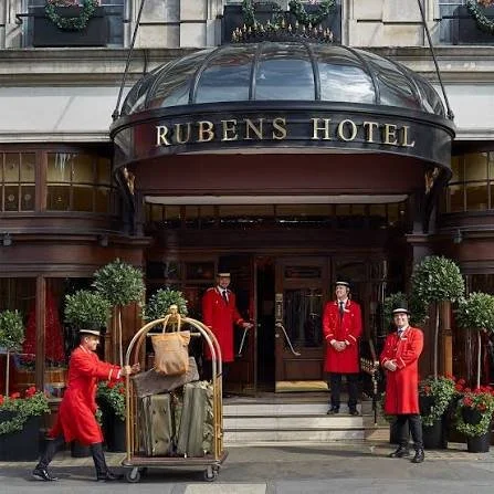 Hotel entrance with a glass dome canopy labeled 'RUBENS HOTEL'. Four bellhops in red uniforms are standing or handling luggage in front of the entrance, which is decorated with potted plants and flowers.
