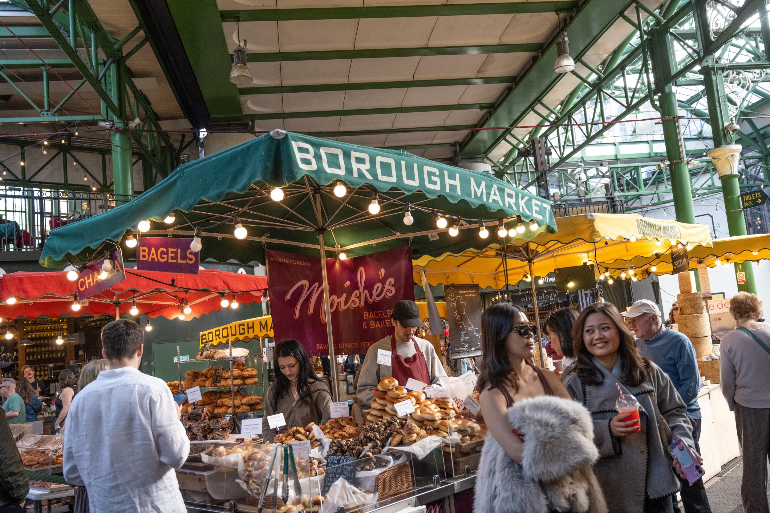 People shopping at Borough Market food stall with baked goods, under green and yellow canopies, inside a large indoor market with green metal framework, signs for bagels and cheese, and strings of lights, with many customers browsing and purchasing food.