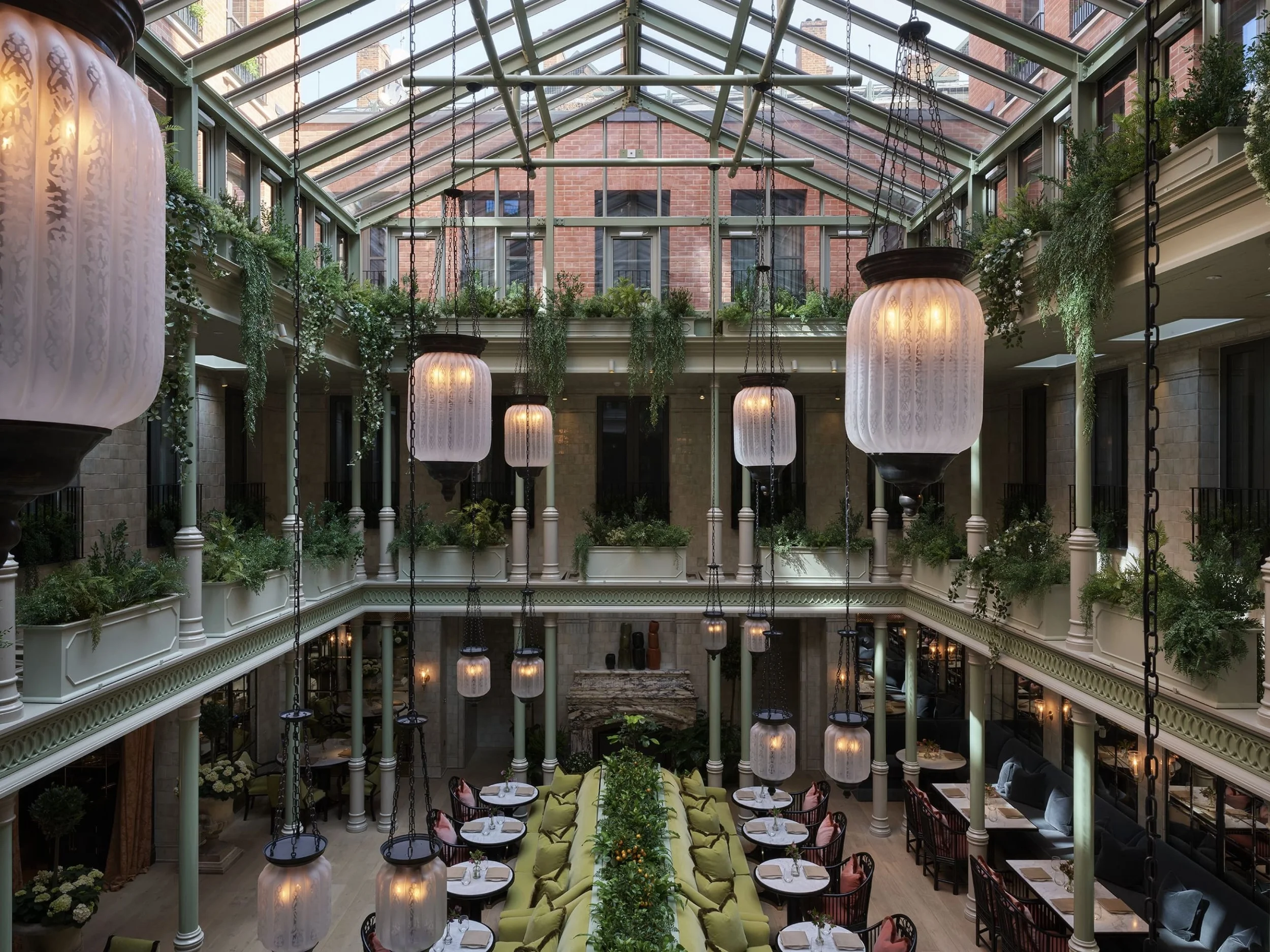 Interior view of a multi-level indoor space with decorative hanging lamps, lush green plants, a glass ceiling, and several dining tables with chairs.
