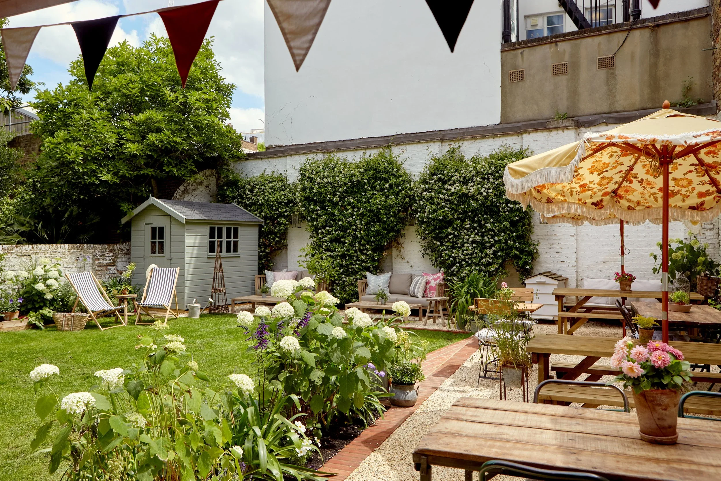 A backyard garden patio with colorful bunting, greenery, a small white shed, outdoor furniture, potted plants, a large yellow floral umbrella, and a brick pathway.