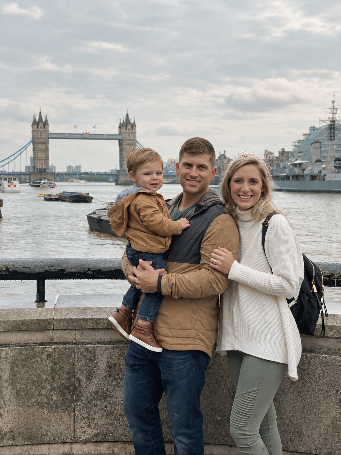 A family of three standing near the River Thames in London, with Tower Bridge and a large navy ship in the background. The family includes a man, woman, and young boy, all smiling at the camera.