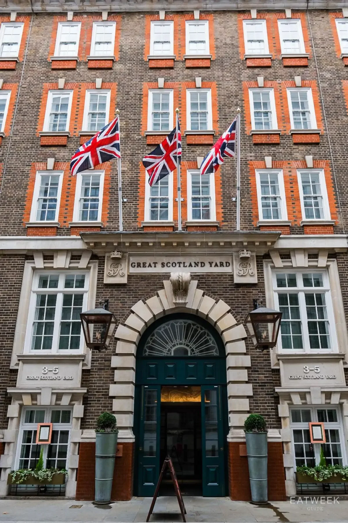 Facade of a historic brick building with sign 'Great Scotland Yard,' three Union Jack flags on flagpoles, and large potted plants flanking the entrance, indicating it is 3-5 Great Scotland Yard.