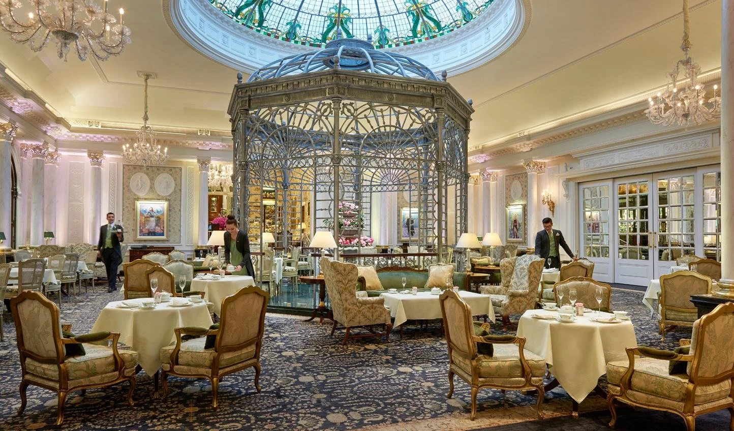 Elegant restaurant interior with round tables covered in white tablecloths, ornate armchairs, large crystal chandeliers, and a glass-domed structure in the center, with waitstaff attending to guests.