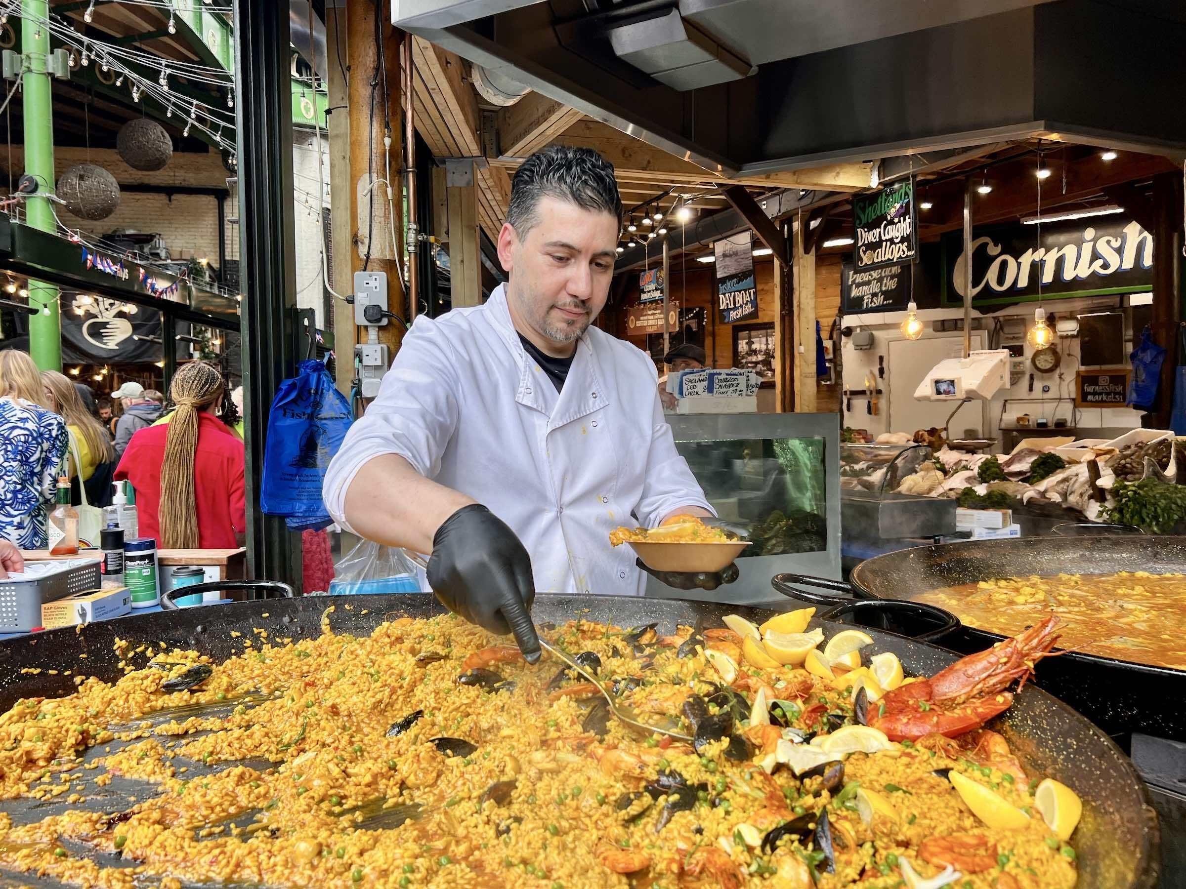 A man in a white chef's coat cooks paella with shrimp and lemon slices in a busy marketplace or food stall, with a crowd and food signs in the background.