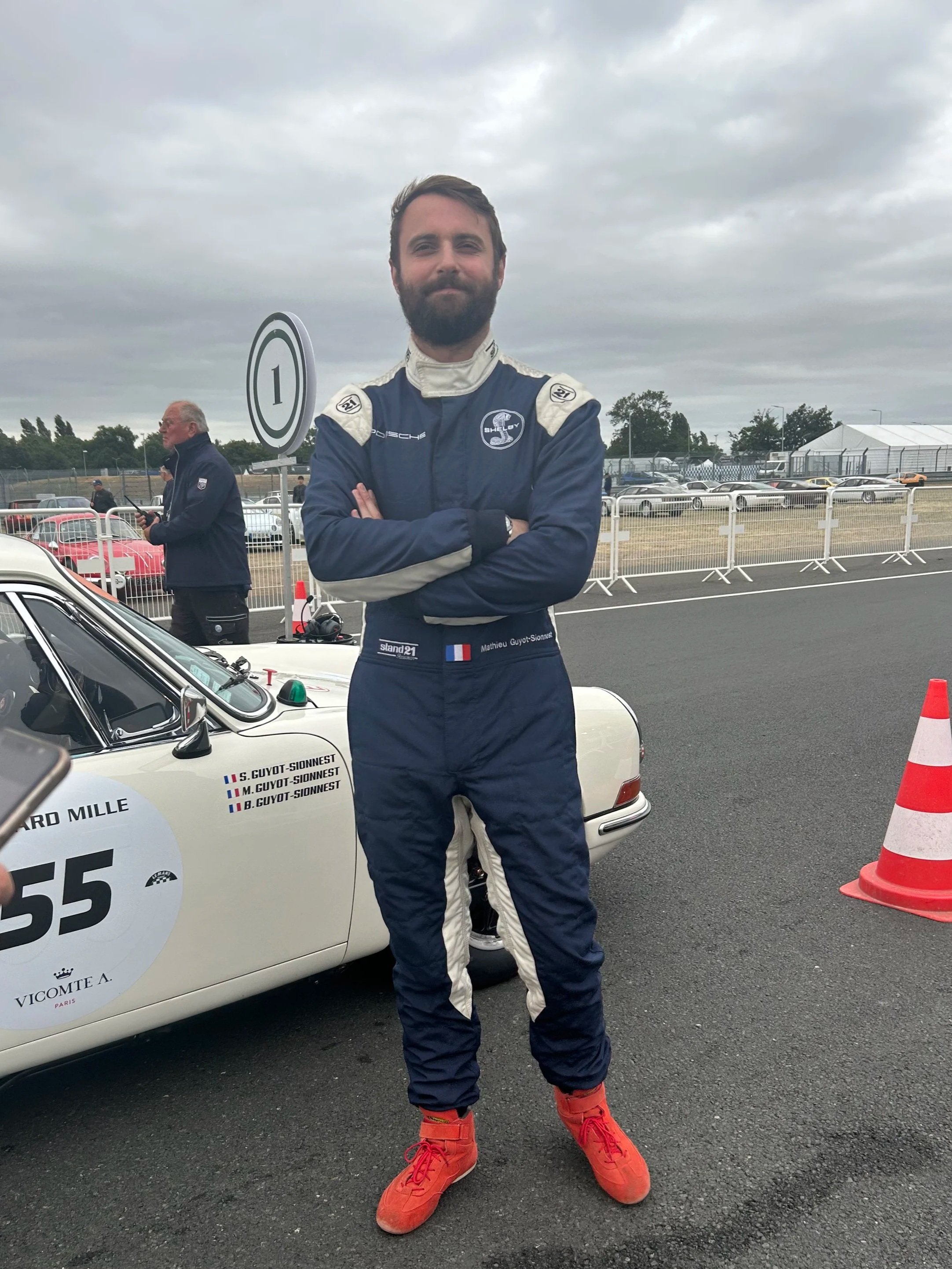 A race car driver in a racing suit and red shoes standing with arms crossed next to a white race car marked with the number 55, at a race track with cars, monitors, and a person in the background.