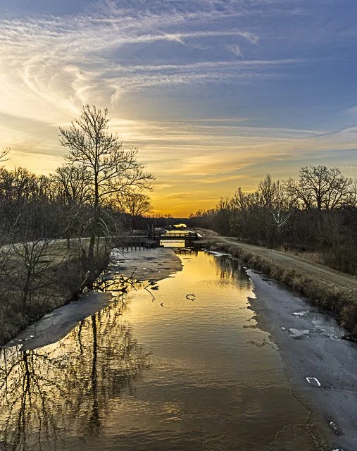 Sunset over a partially frozen river with leafless trees along the banks and a dirt road to the right.
