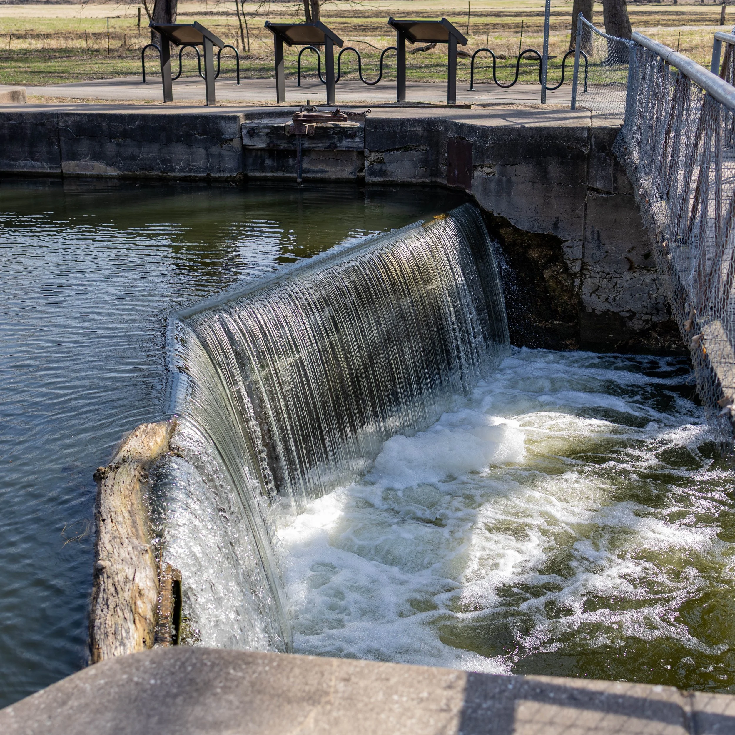 A small dam with water flowing over it, surrounded by a metal fence and walkways, in a park with grassy fields and trees in the background.
