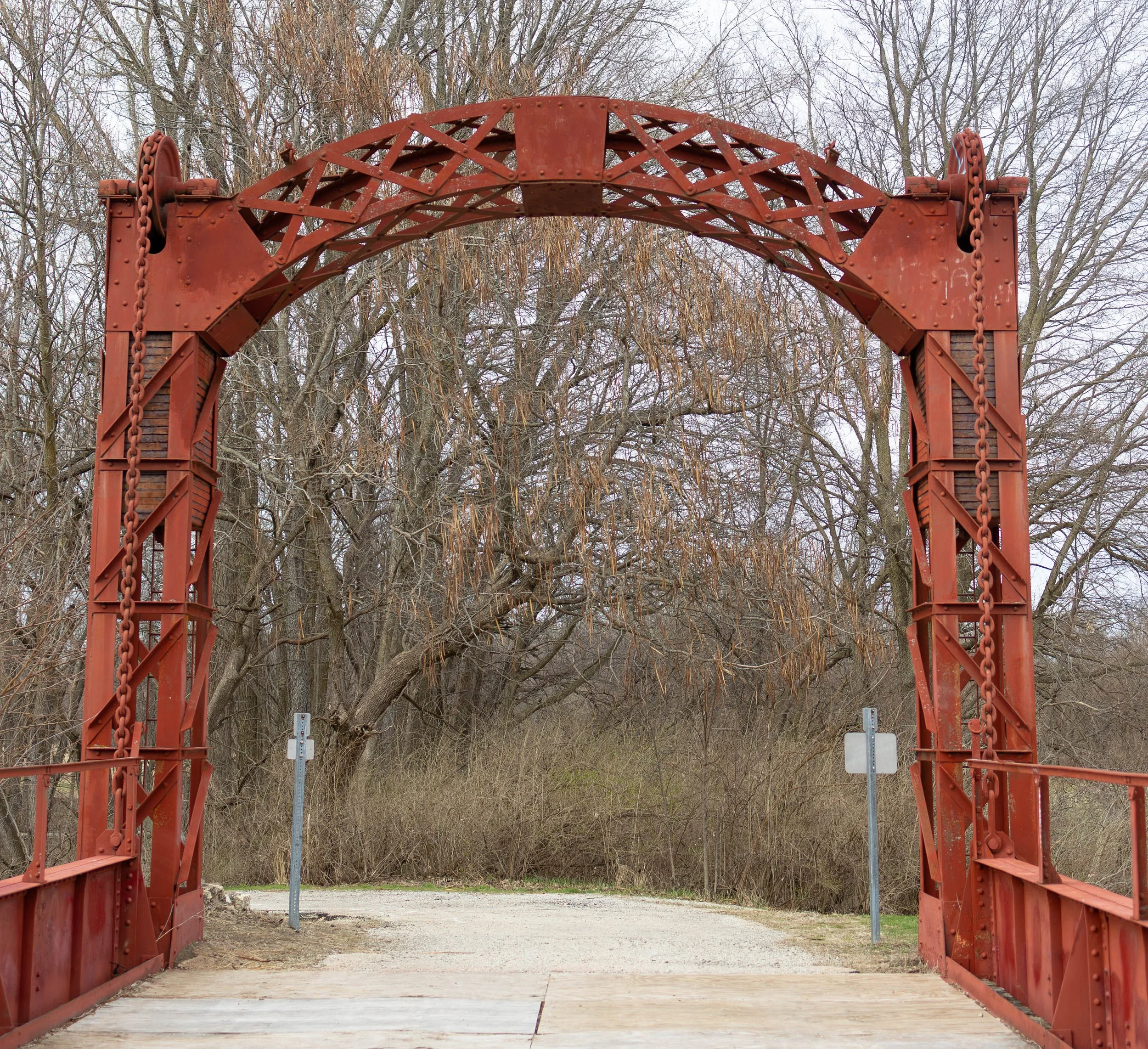Old red metal arch bridge over a gravel path, surrounded by leafless trees in a natural setting.